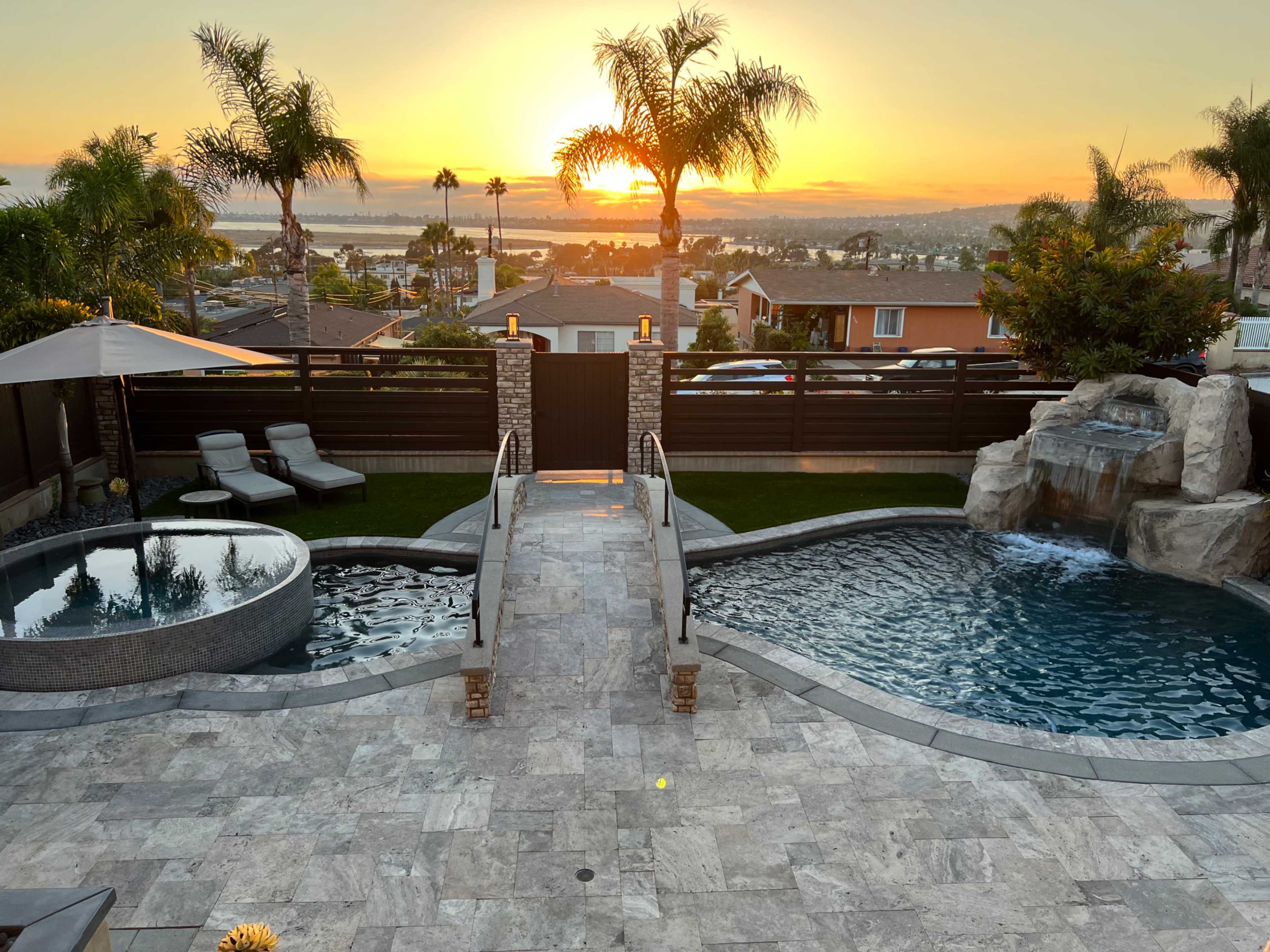 A backyard with a spa, swimming pool, and a sunset backdrop, framed by palm trees and a landscaped garden.