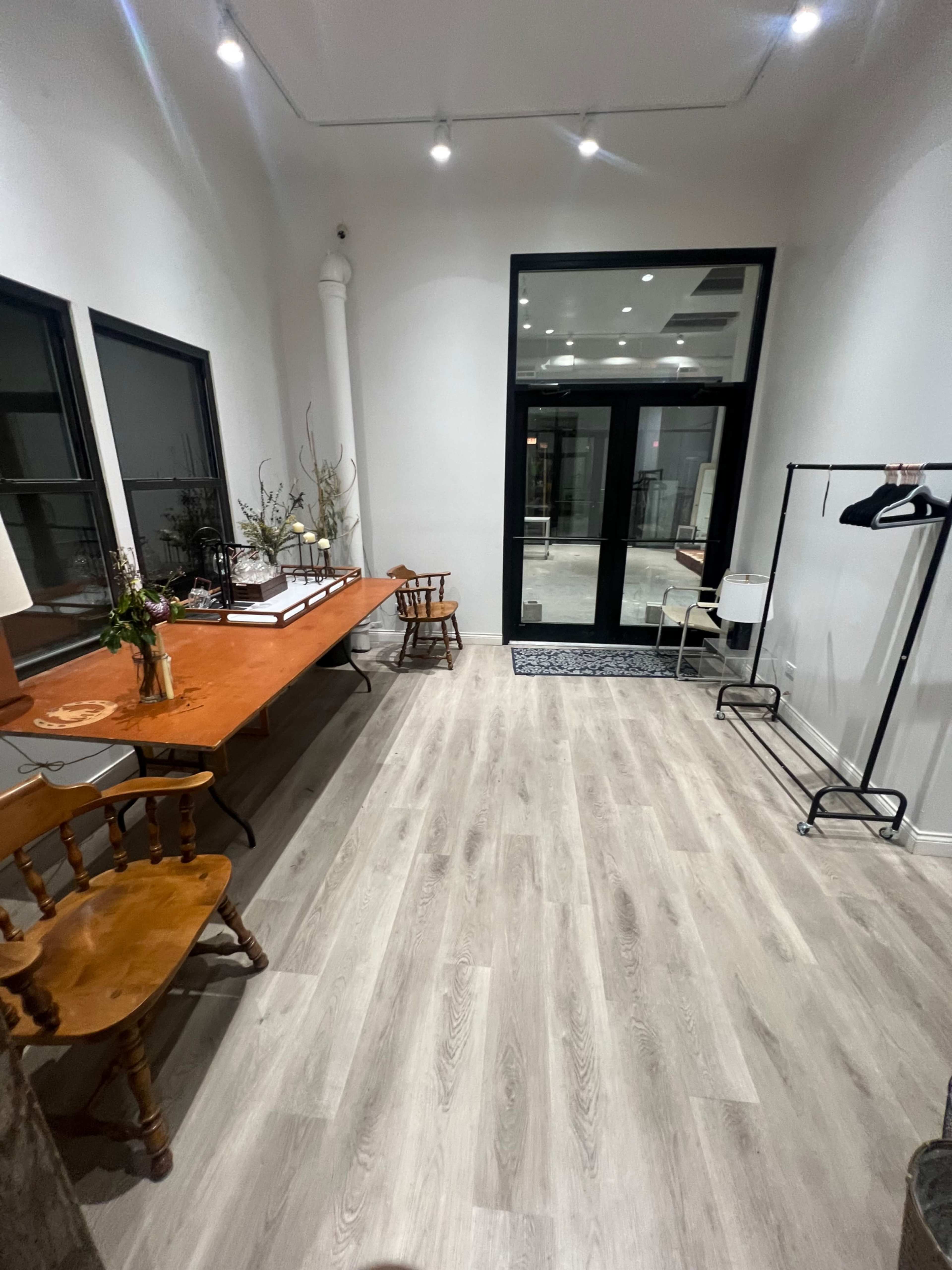 The image shows a spacious interior with wooden flooring, featuring a long orange table, a few chairs, and a clothing rack against a wall.