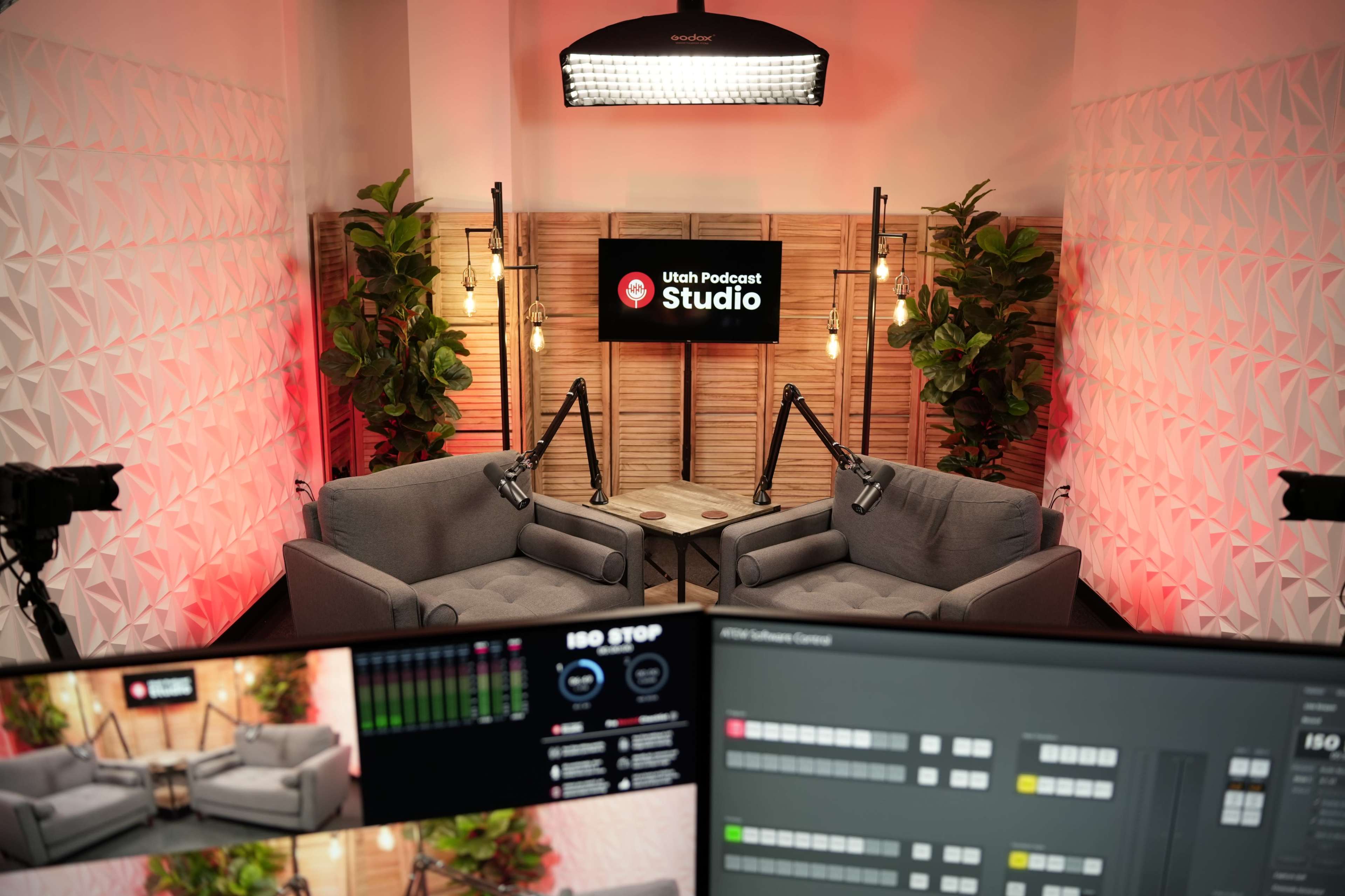 The image shows a podcast studio with two gray sofas, camera equipment, and a screen displaying "Utah Podcast Studio," surrounded by decorative plants and studio lighting.