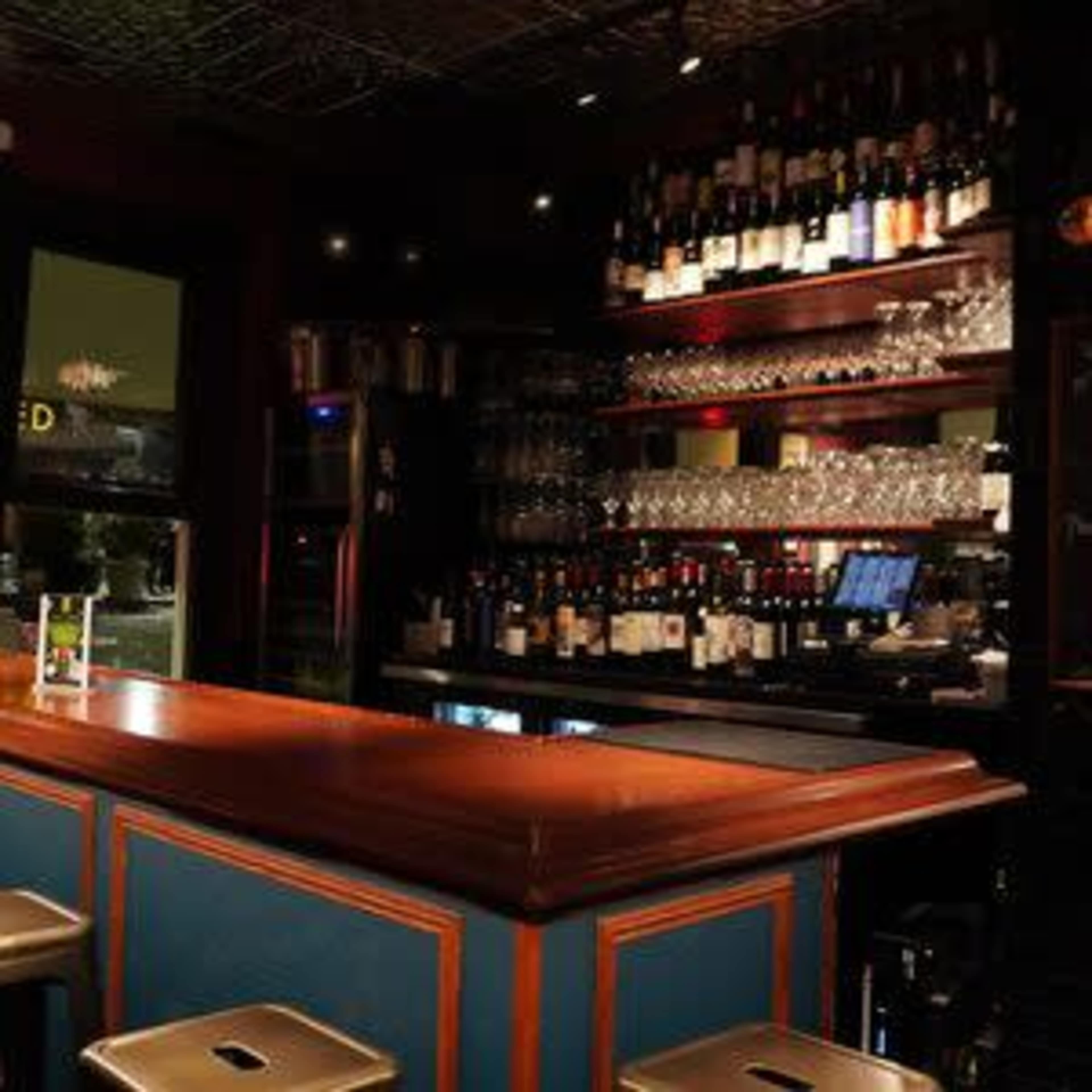 The image shows a well-organized bar with a polished wooden counter, shelves filled with various bottles of wine and liquor, and glasses neatly arranged.