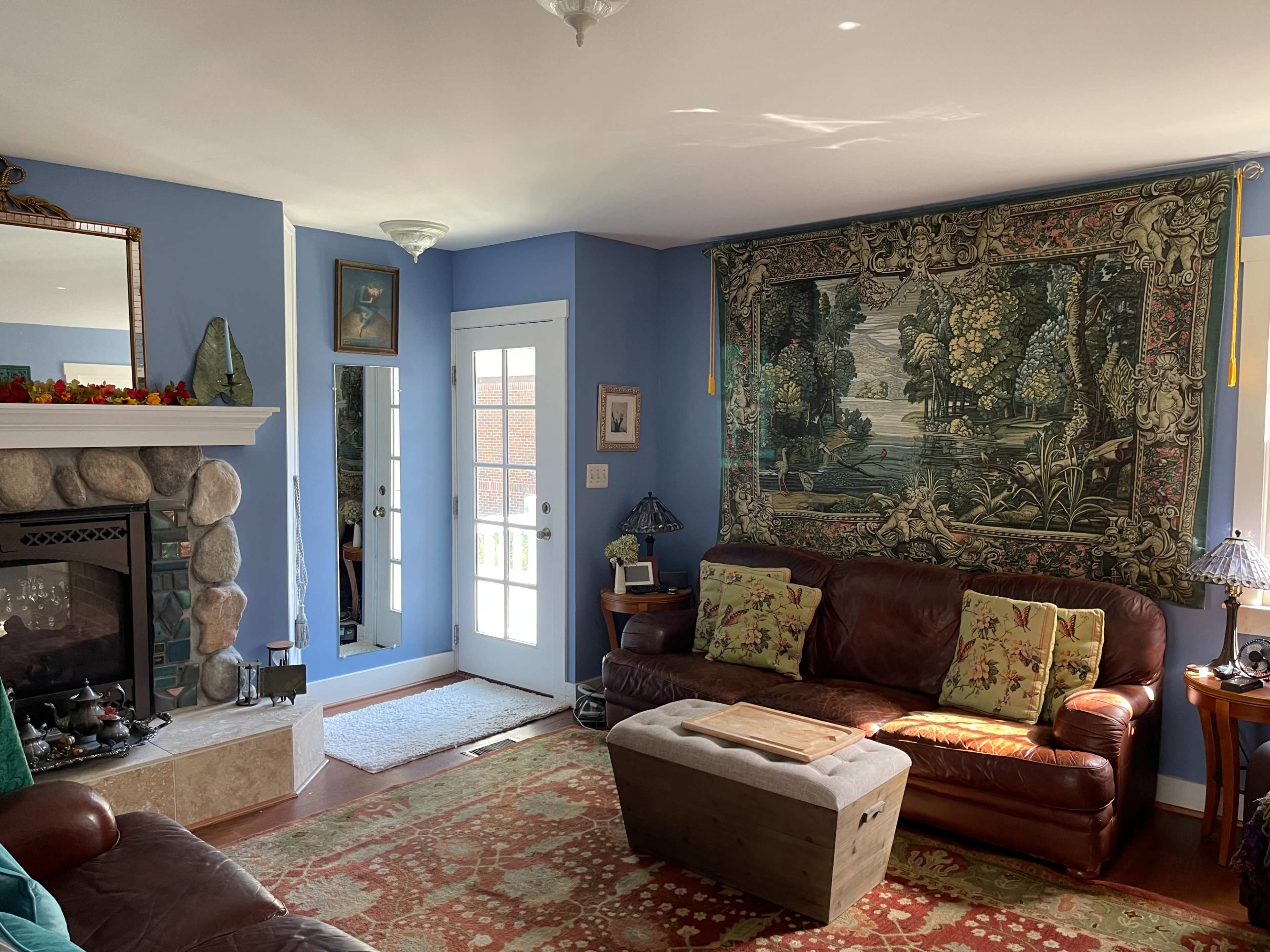 A living room with blue walls, a stone fireplace, a large tapestry on one side, and leather couches arranged around a wooden coffee table.