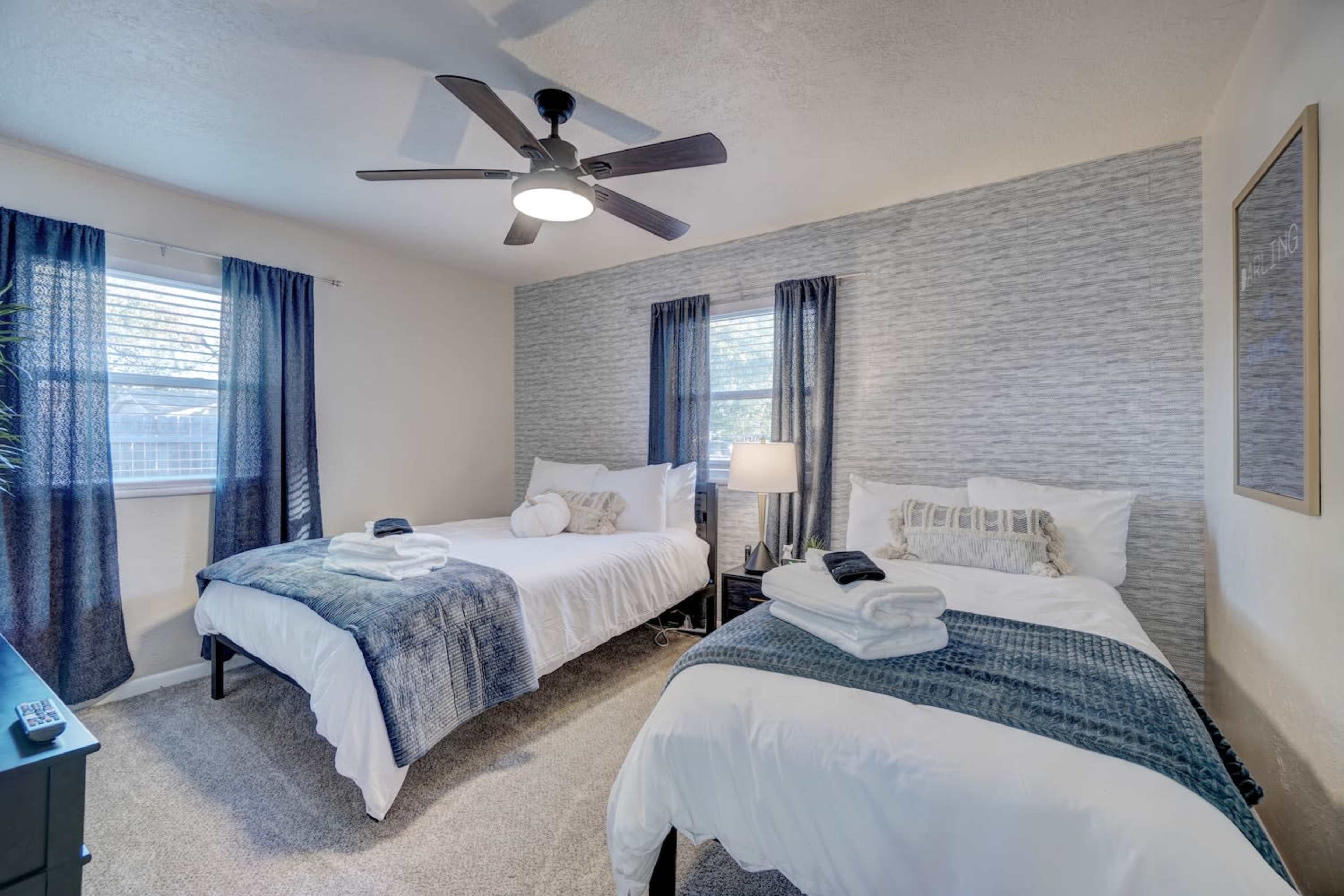 A bedroom with two twin beds, a ceiling fan, and two windows with blue curtains.