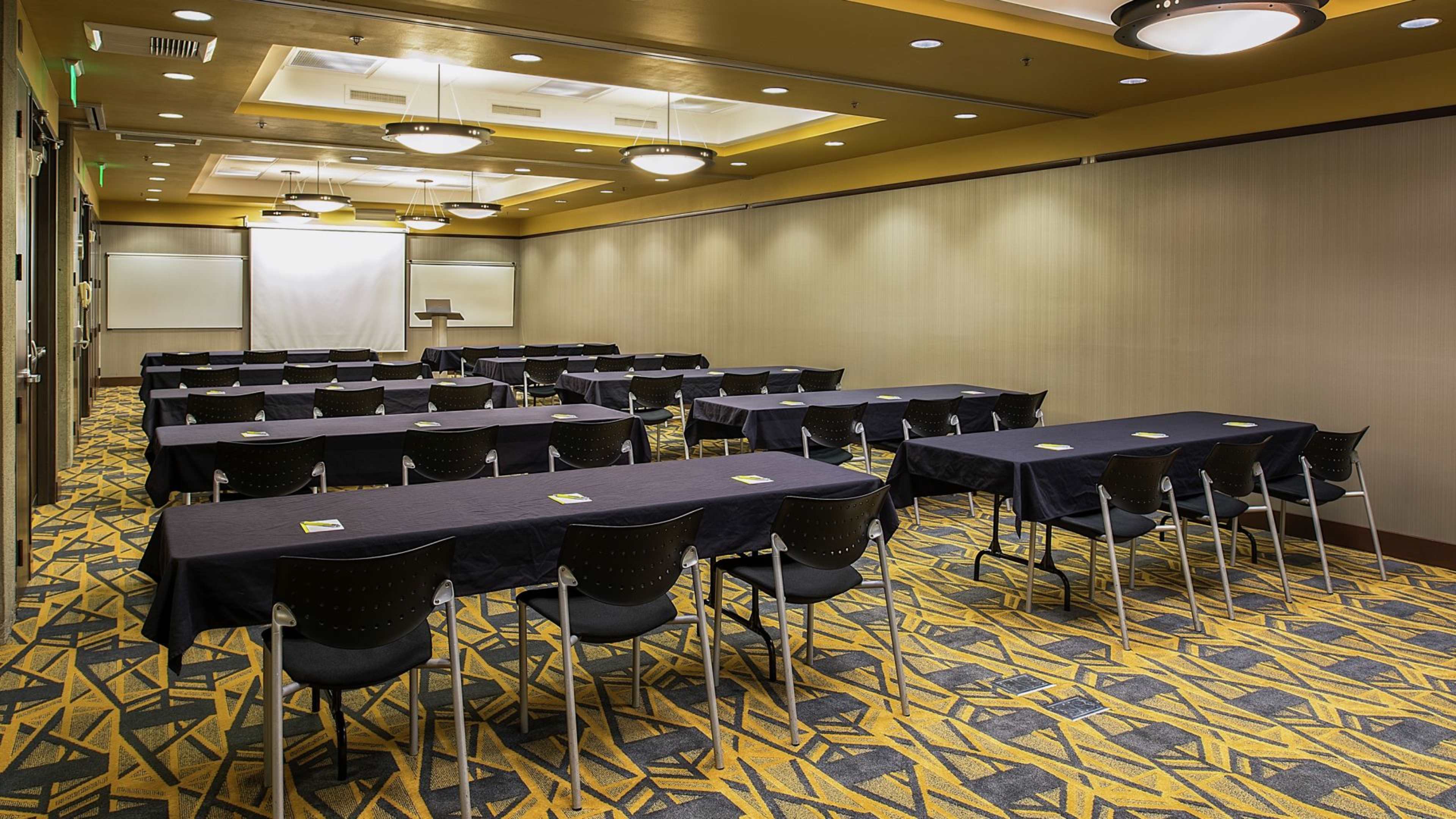 A conference room with multiple tables arranged in rows, each covered with black tablecloths and facing a projection screen.