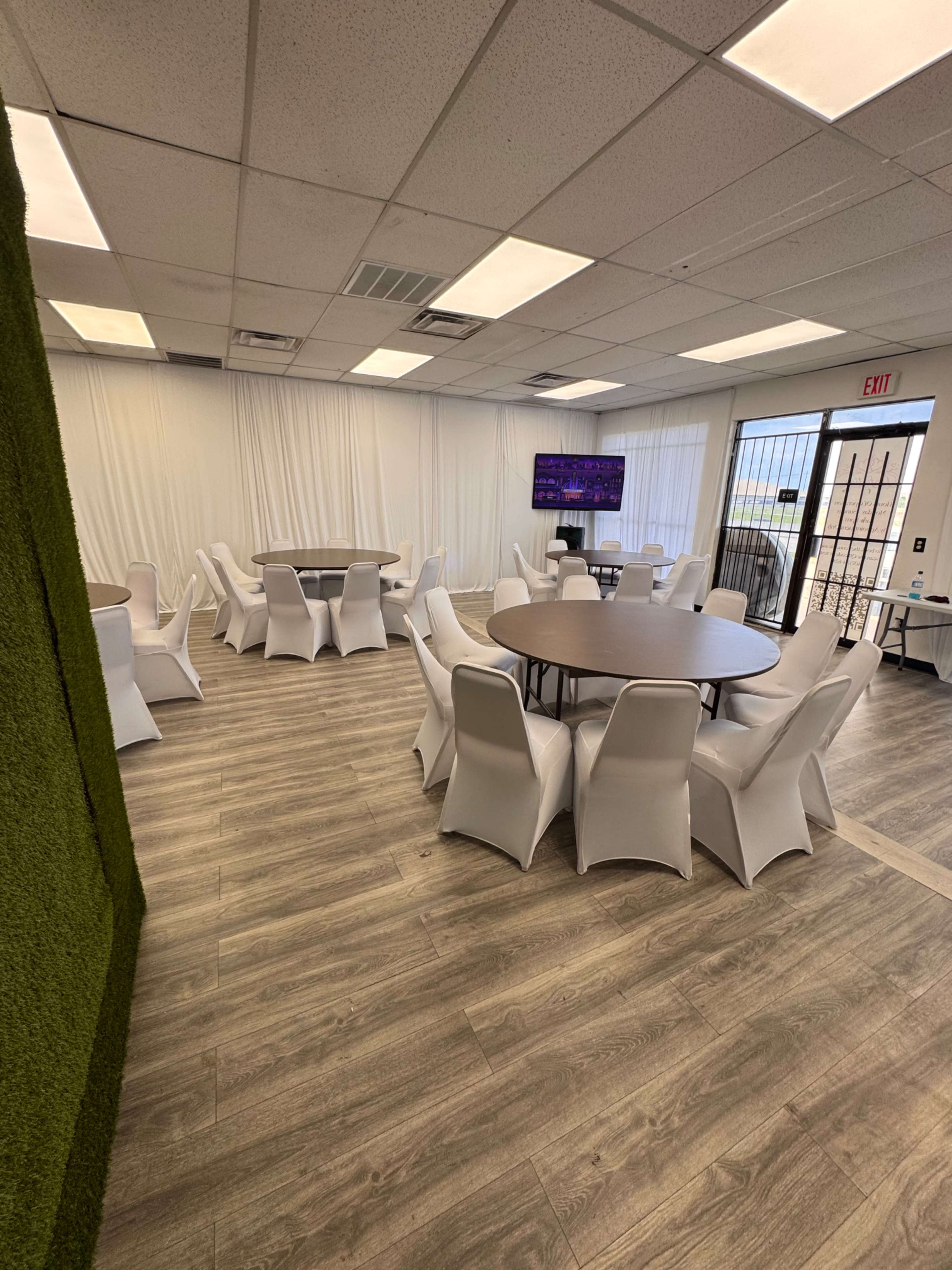 The image shows a spacious event room with multiple round tables covered in white tablecloths and white chairs, featuring a green wall and a large screen on one side.