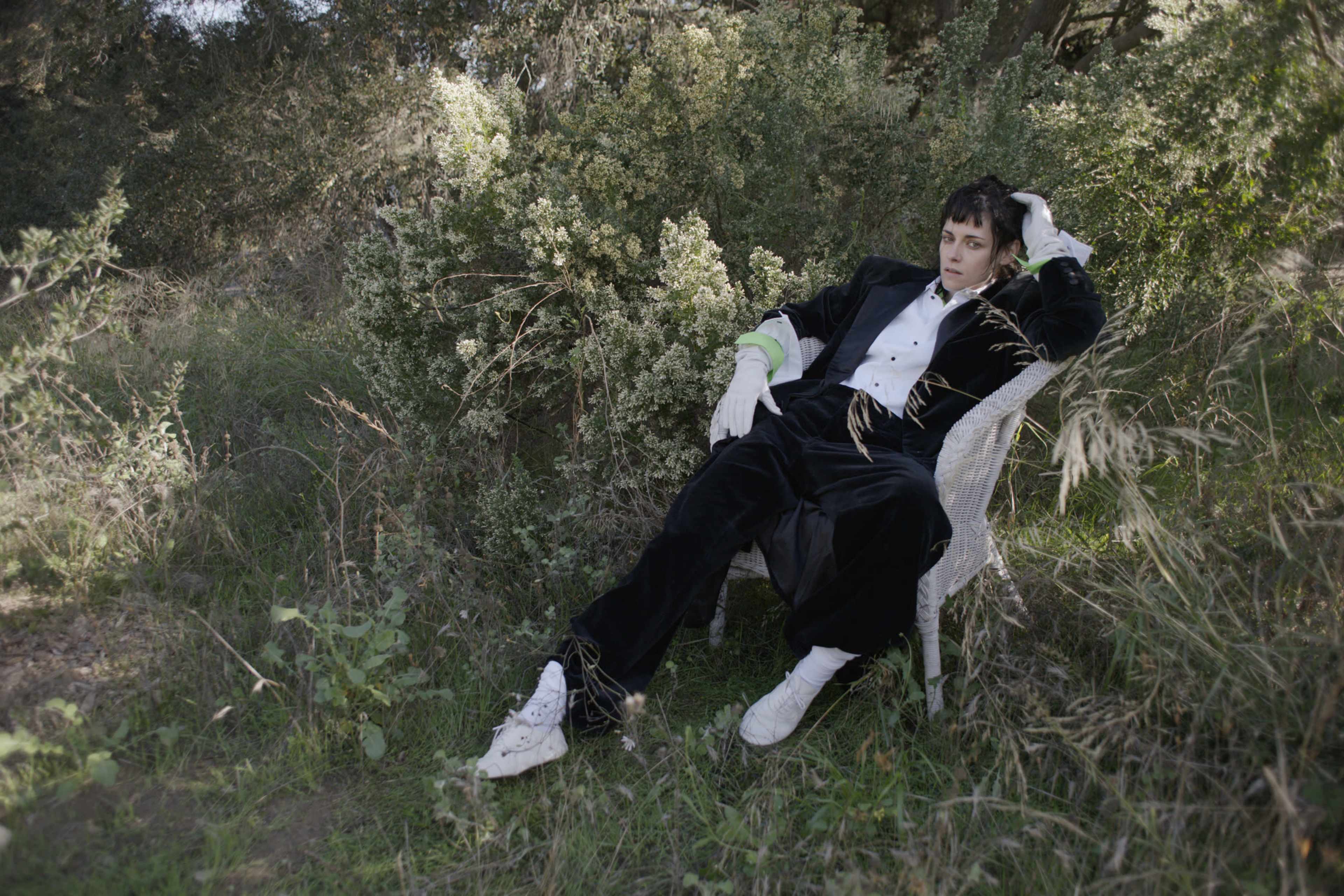 A person wearing a tuxedo sits casually in a white chair among greenery.