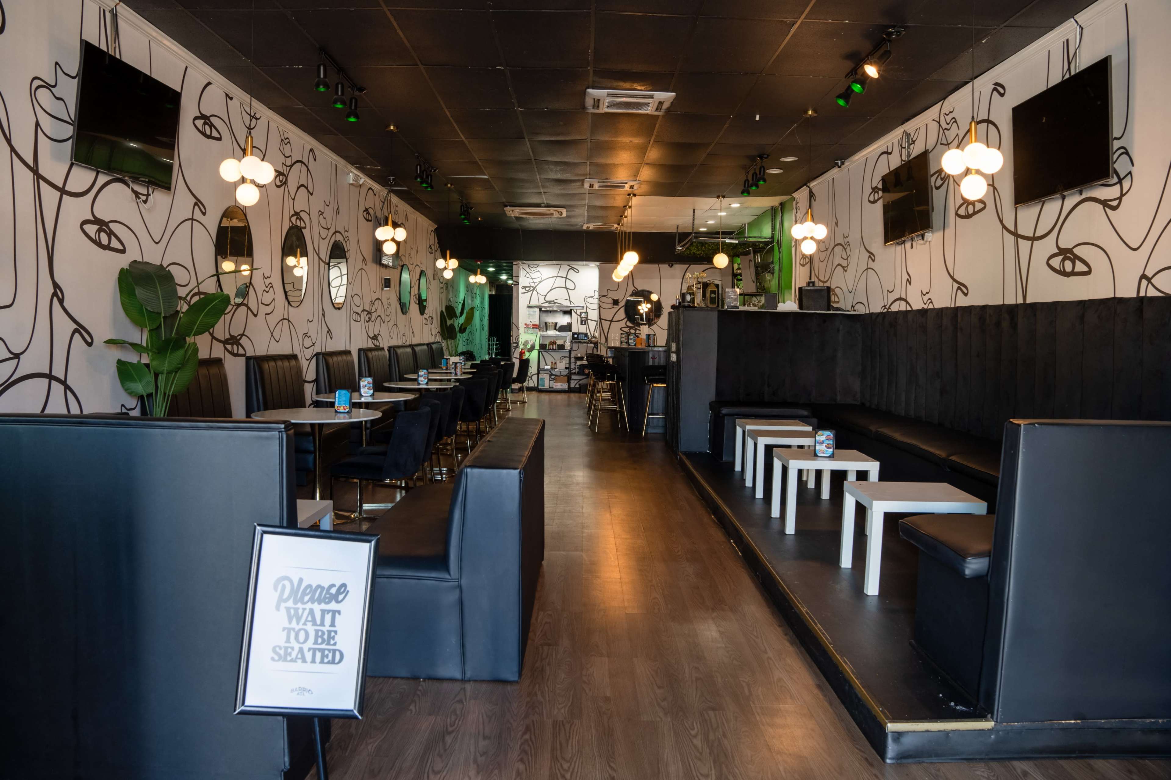 The image shows the interior of a modern café with black and white furnishings, circular light fixtures, and a sign instructing customers to wait to be seated.