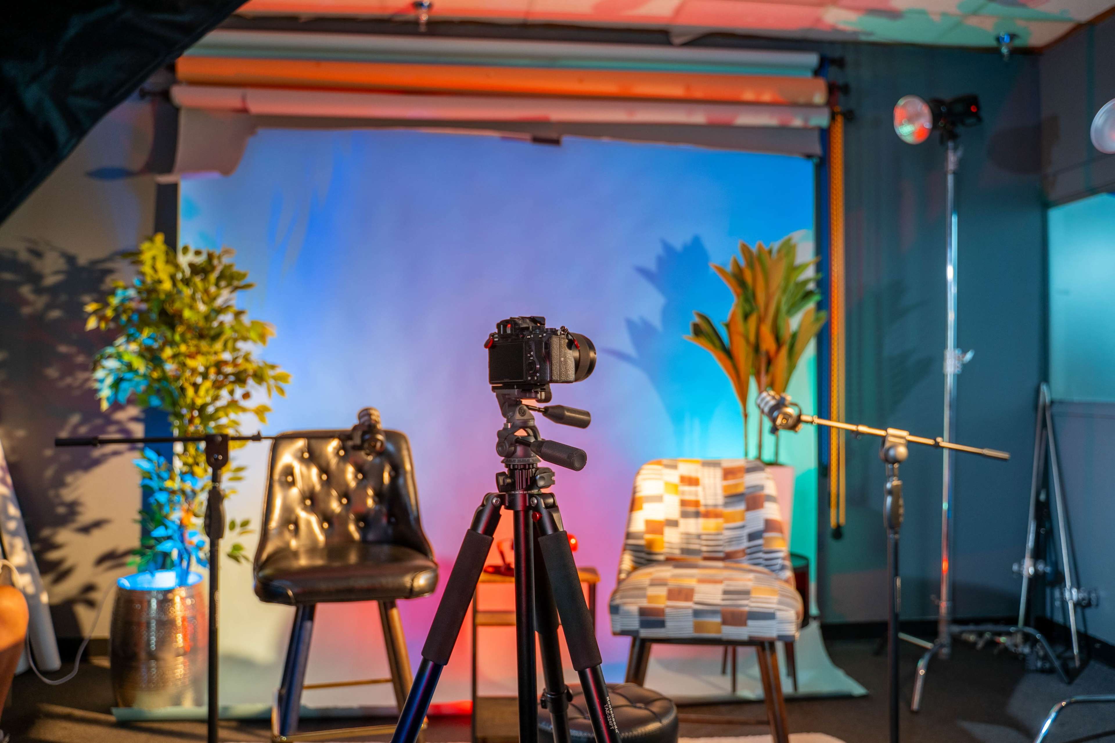 The image shows a photography setup with a camera on a tripod facing two chairs, a potted plant, and colorful backdrop lighting.