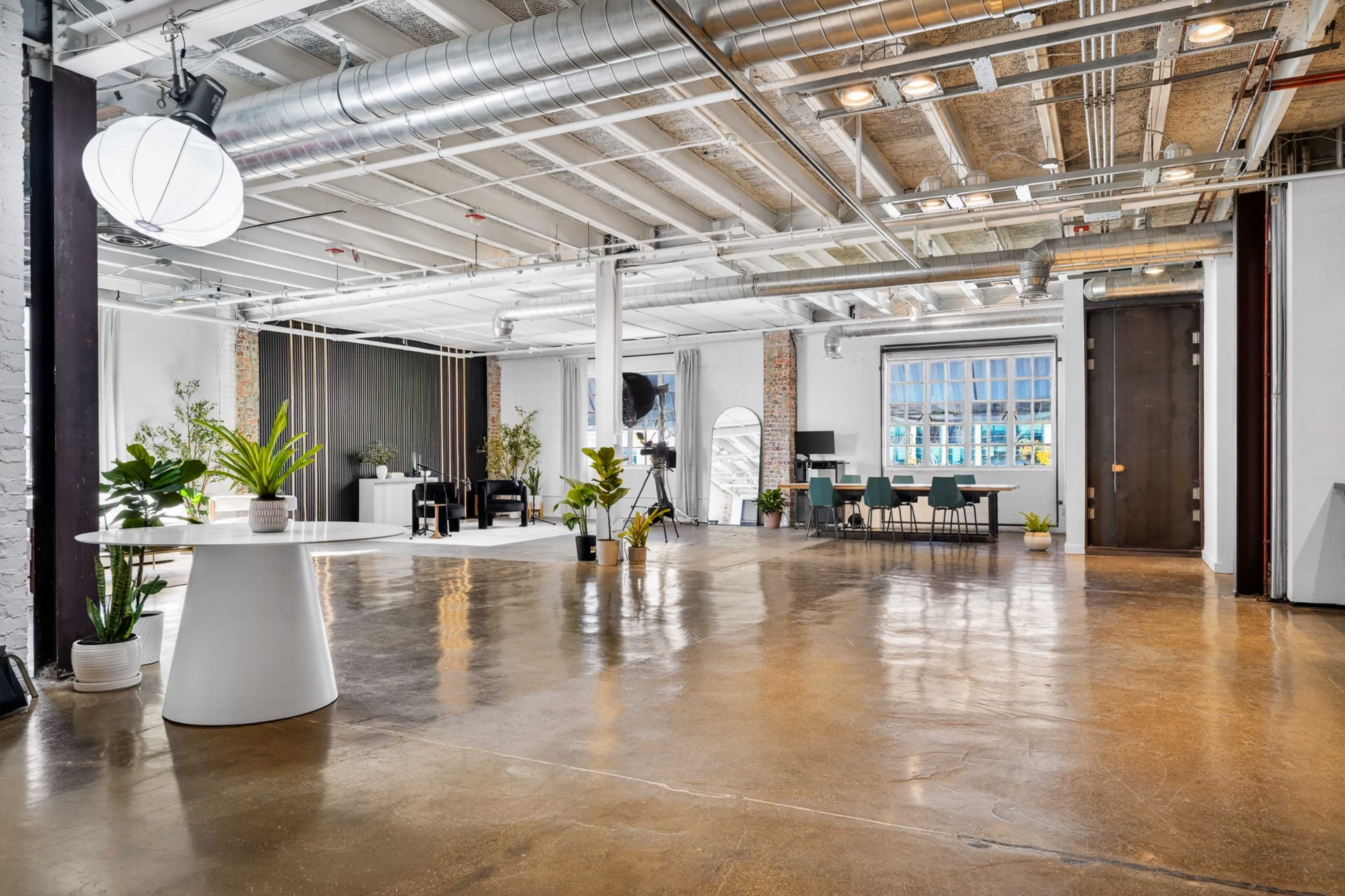 The image features a spacious, modern interior with polished concrete floors, tall ceilings, and a mix of plant decorations and furniture arranged for a workspace or event area.