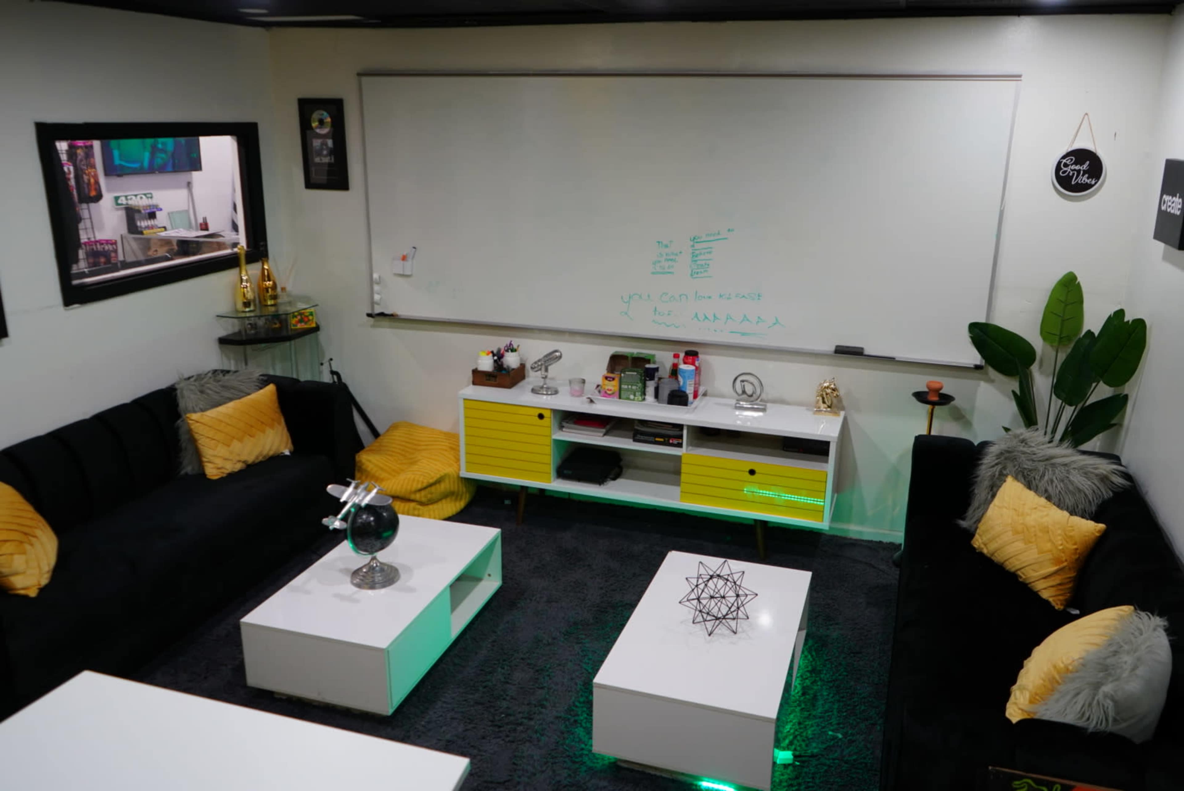 A modern meeting space with black sofas, a whiteboard, a yellow-accented media console, and decorative plants.