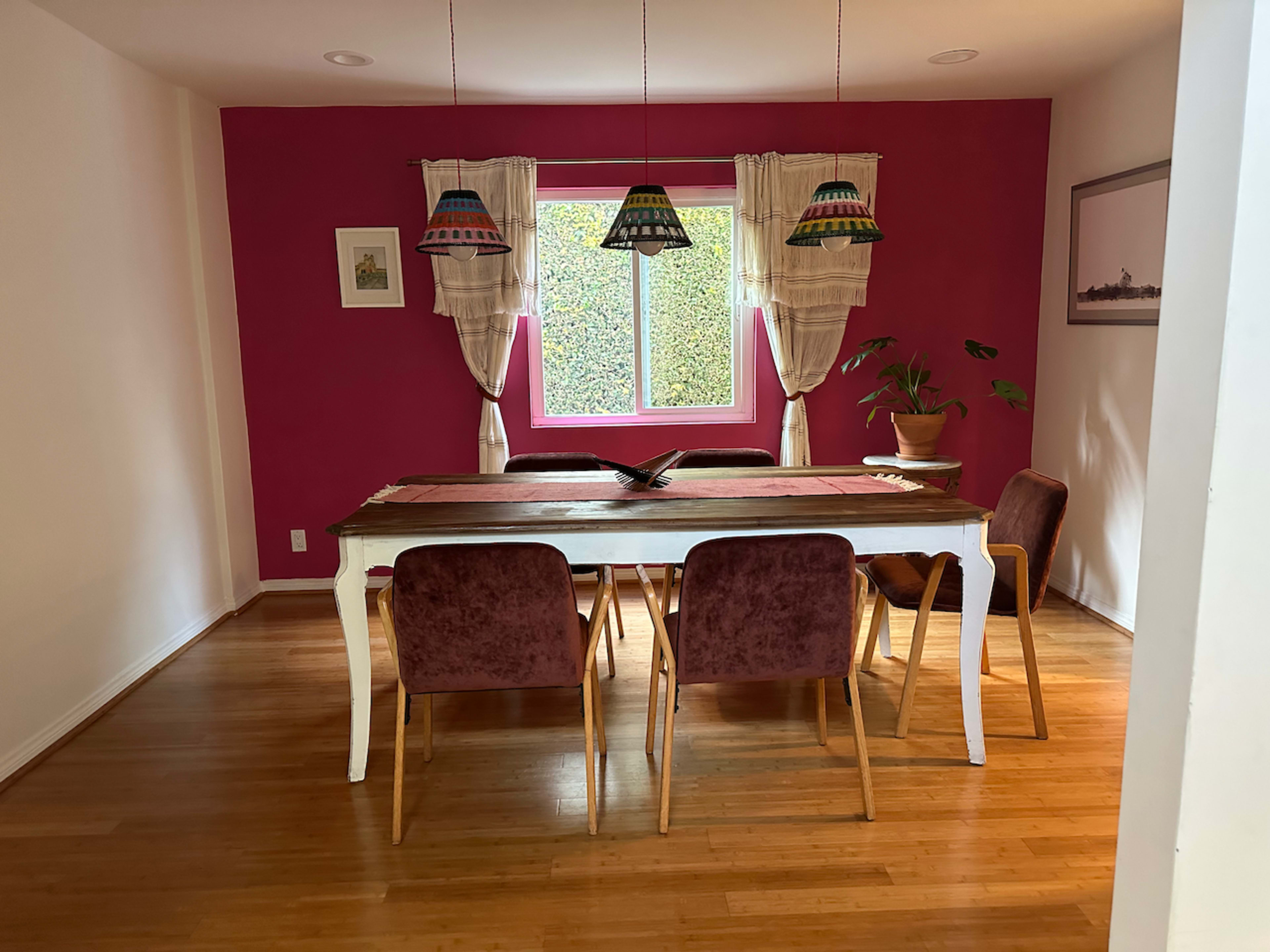 A dining room features a wooden table surrounded by six chairs, with colorful pendant lights hanging above and a window covered by sheer curtains.