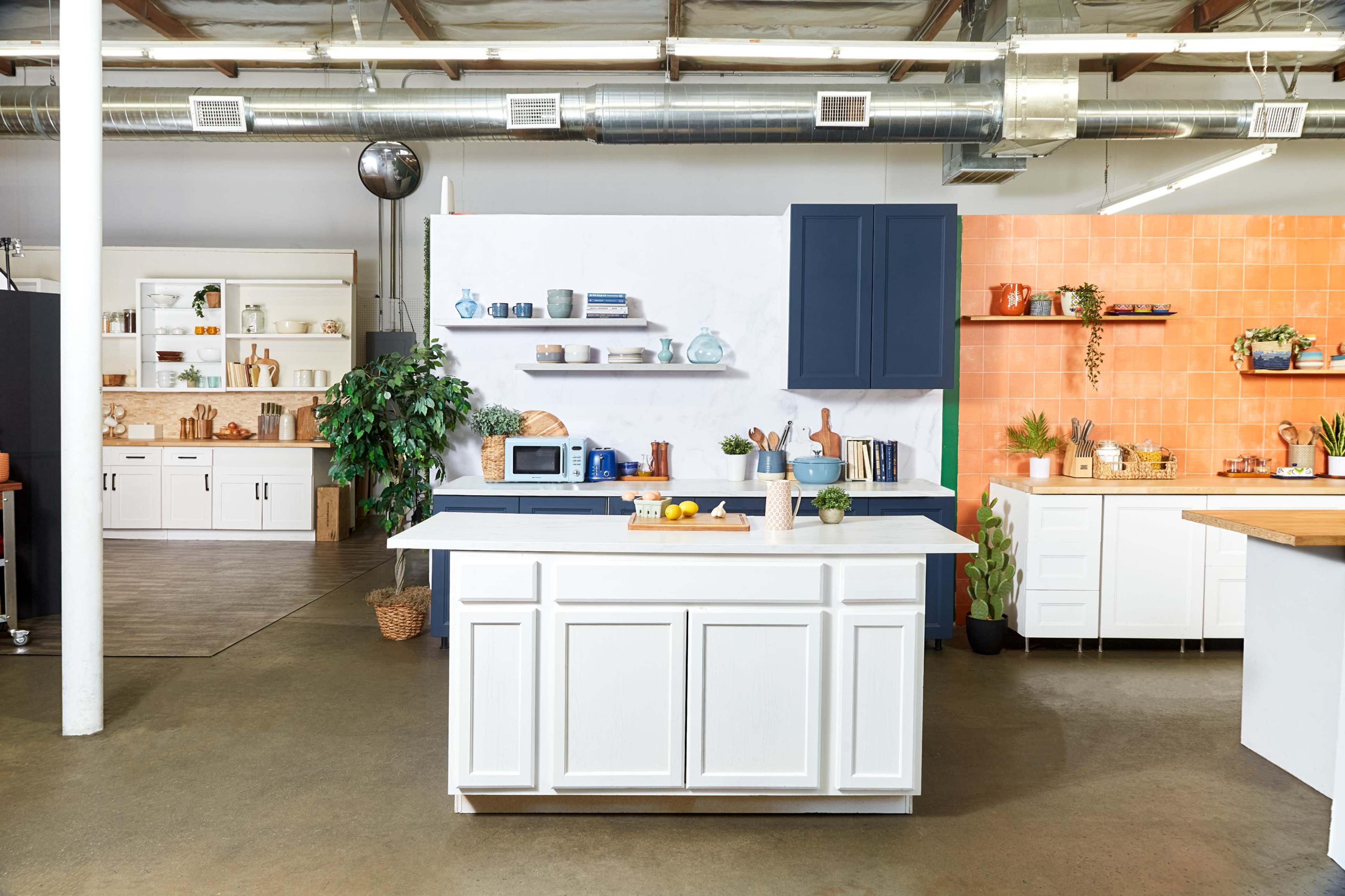 A modern kitchen space featuring a white island with a microwave, surrounded by various shelves and vibrant orange and blue accent walls.