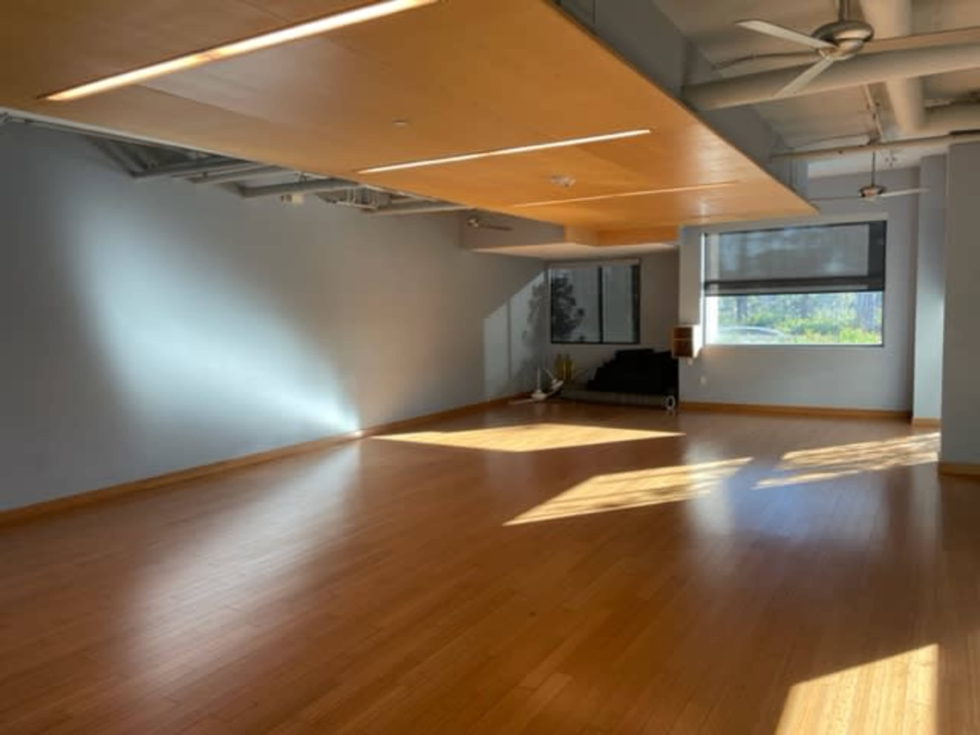 A bright, spacious room with hardwood flooring, large windows, and minimal furnishings, featuring a prominent overhead structure.