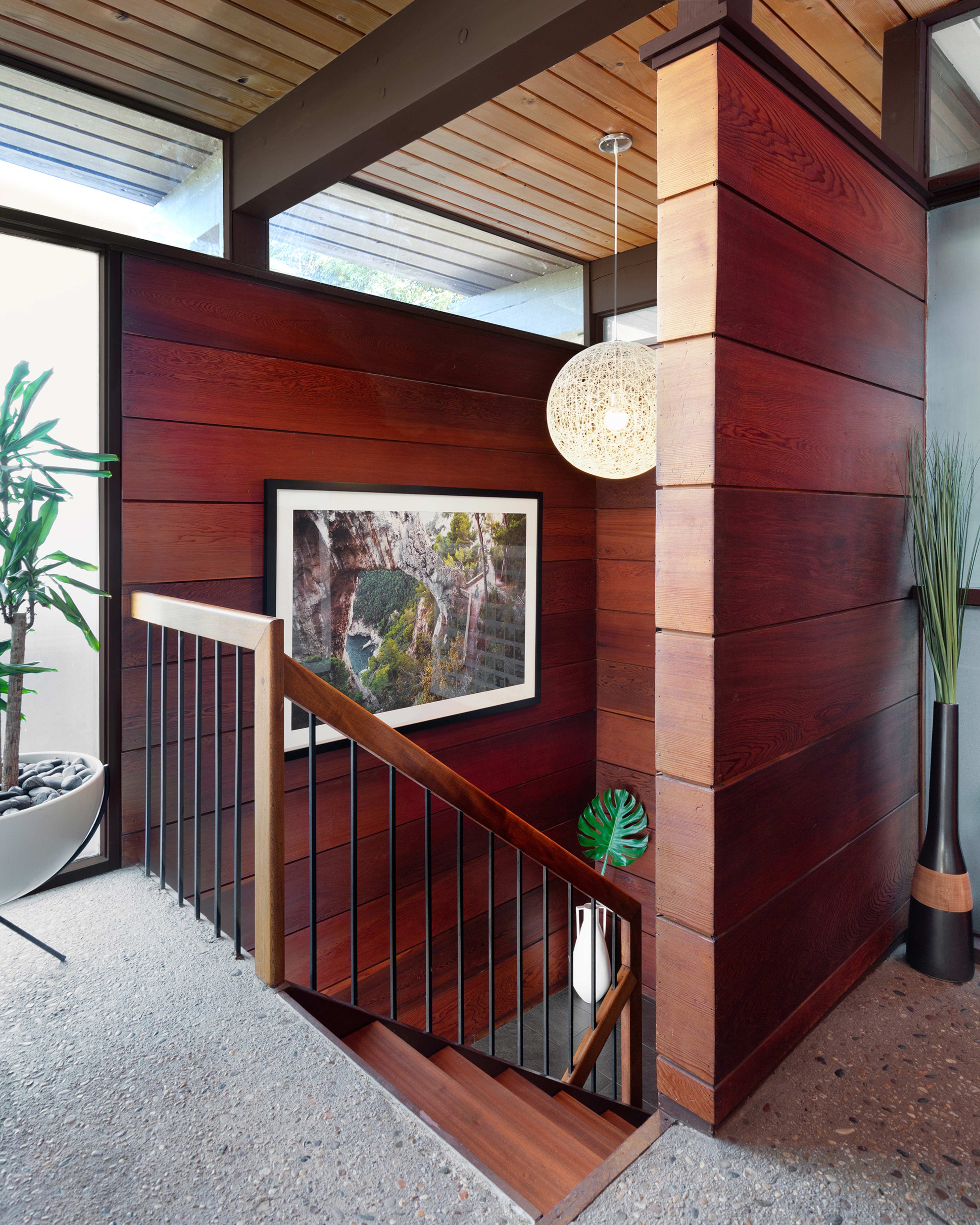 The image shows a modern staircase with wooden paneling and a decorative pendant light, framed by large windows and plants.