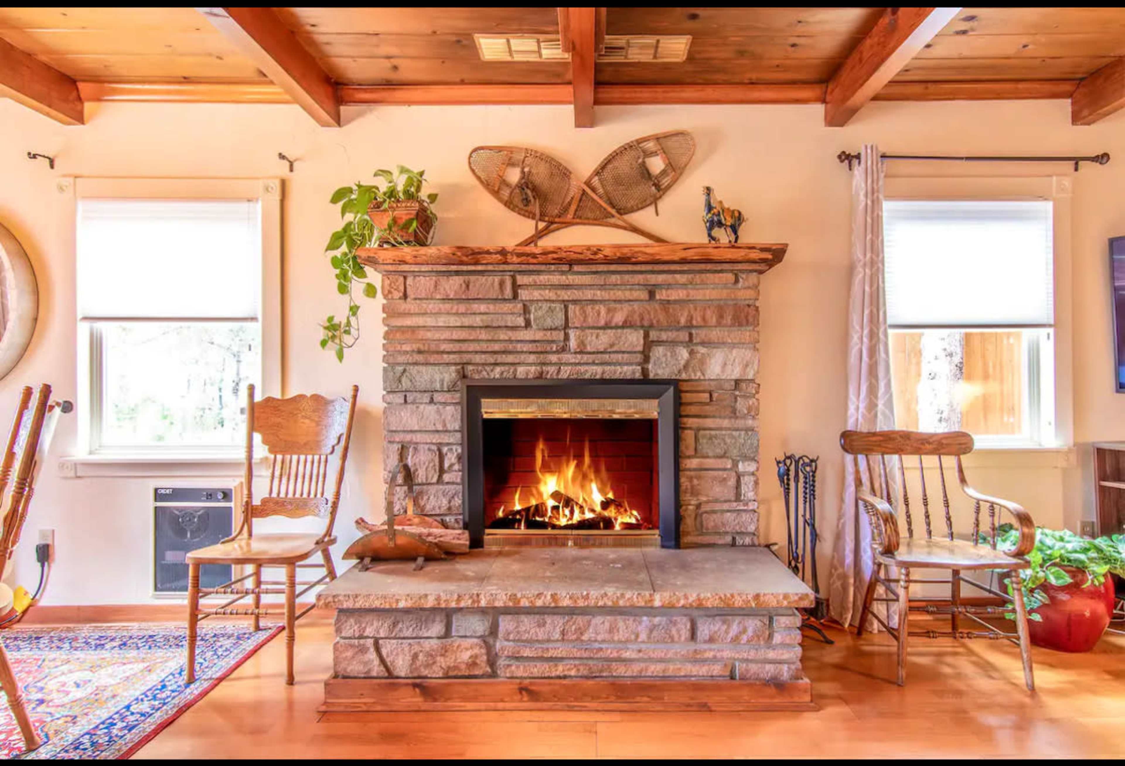 The image shows a cozy living room with a stone fireplace, wooden ceiling beams, and comfortable seating arrangements.
