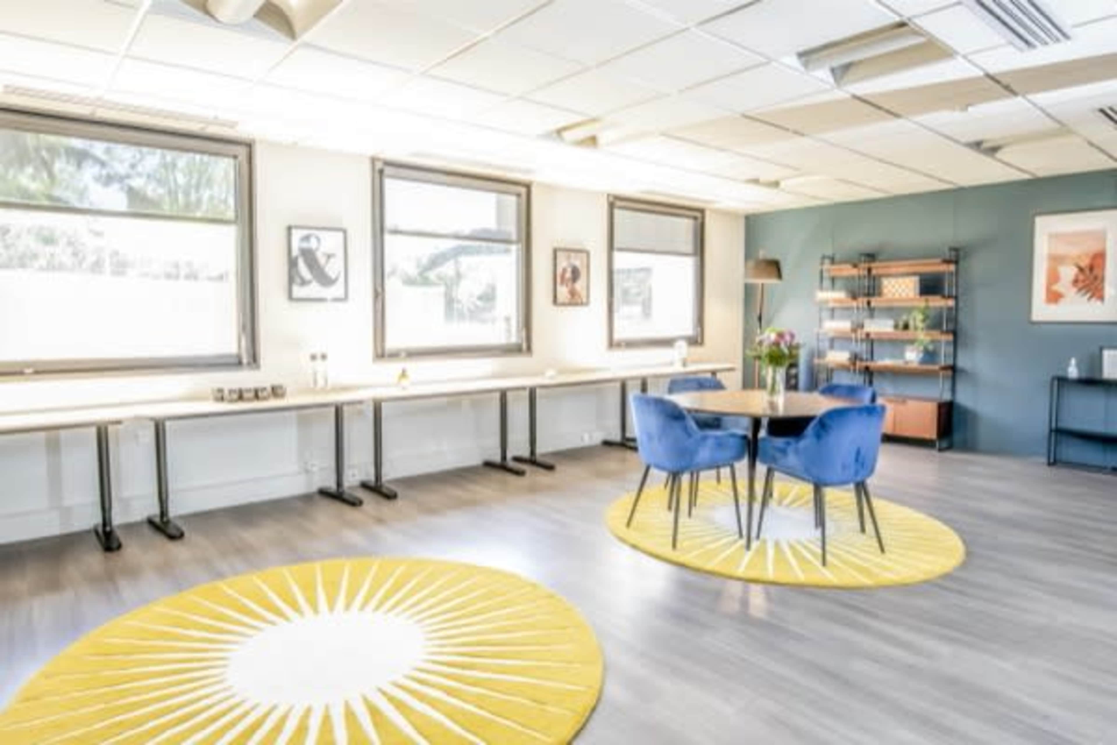 A bright office space featuring two blue chairs around a round table, with yellow sunburst rugs on a wooden floor and open windows allowing natural light.