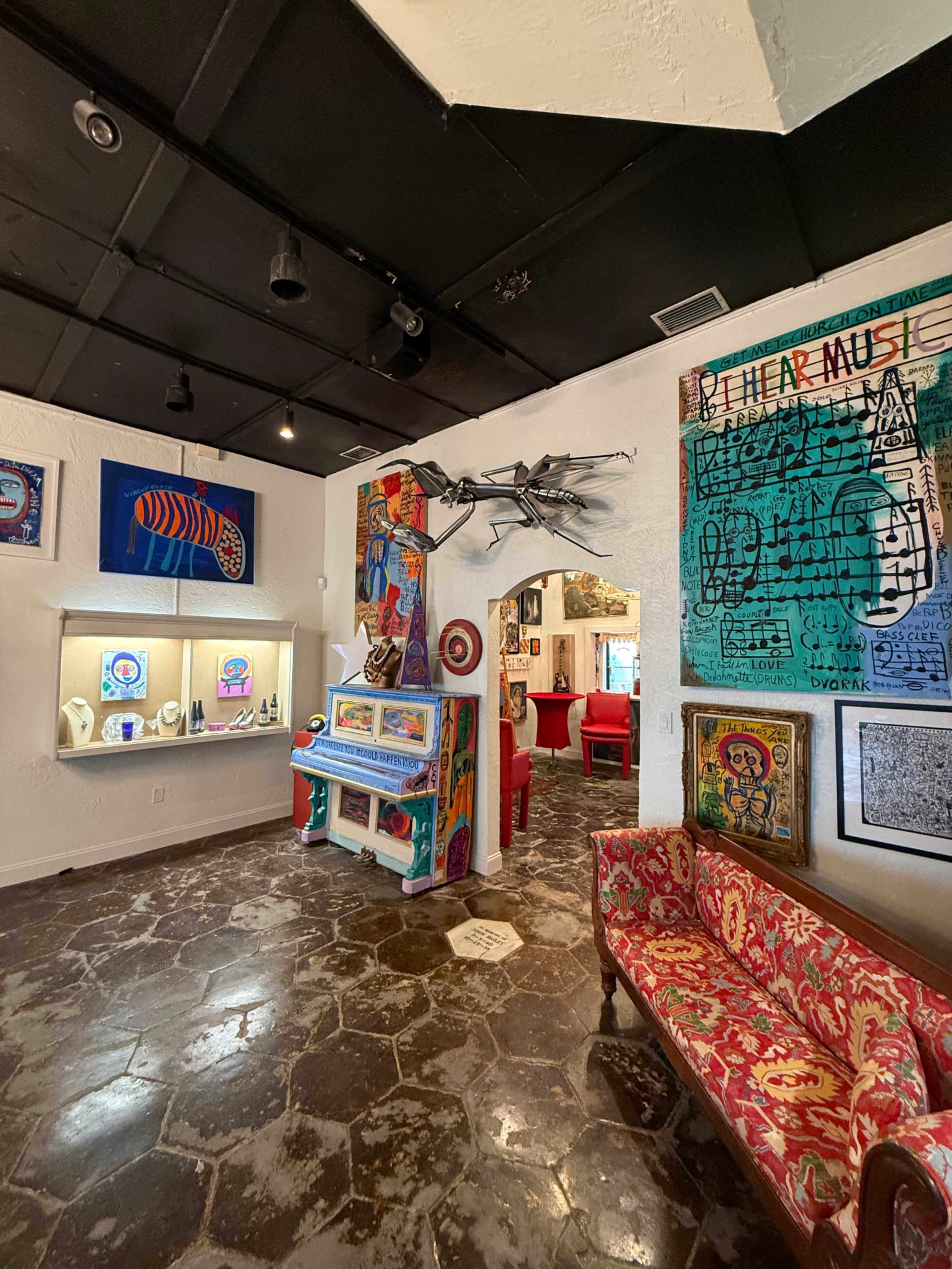 The image shows an art gallery featuring colorful artworks on the walls, a decorative piano in the center, and red upholstered furniture.