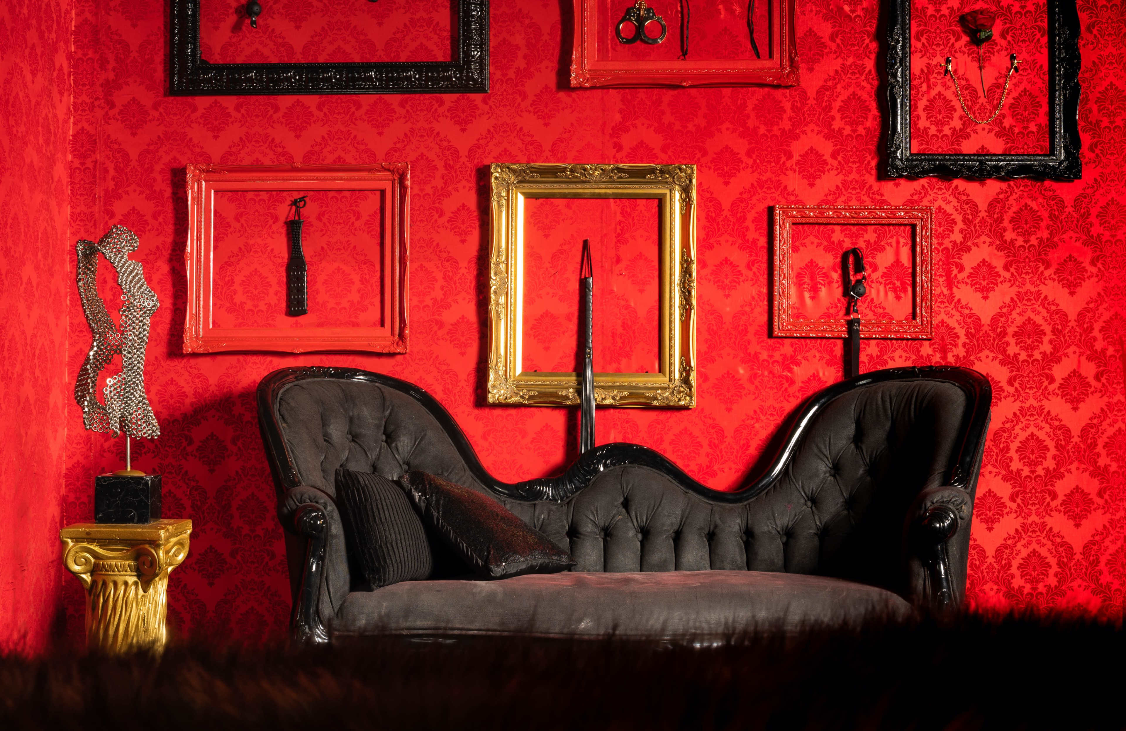 A black velvet couch positioned against a red damask wall adorned with ornate picture frames and decorative sculptures.