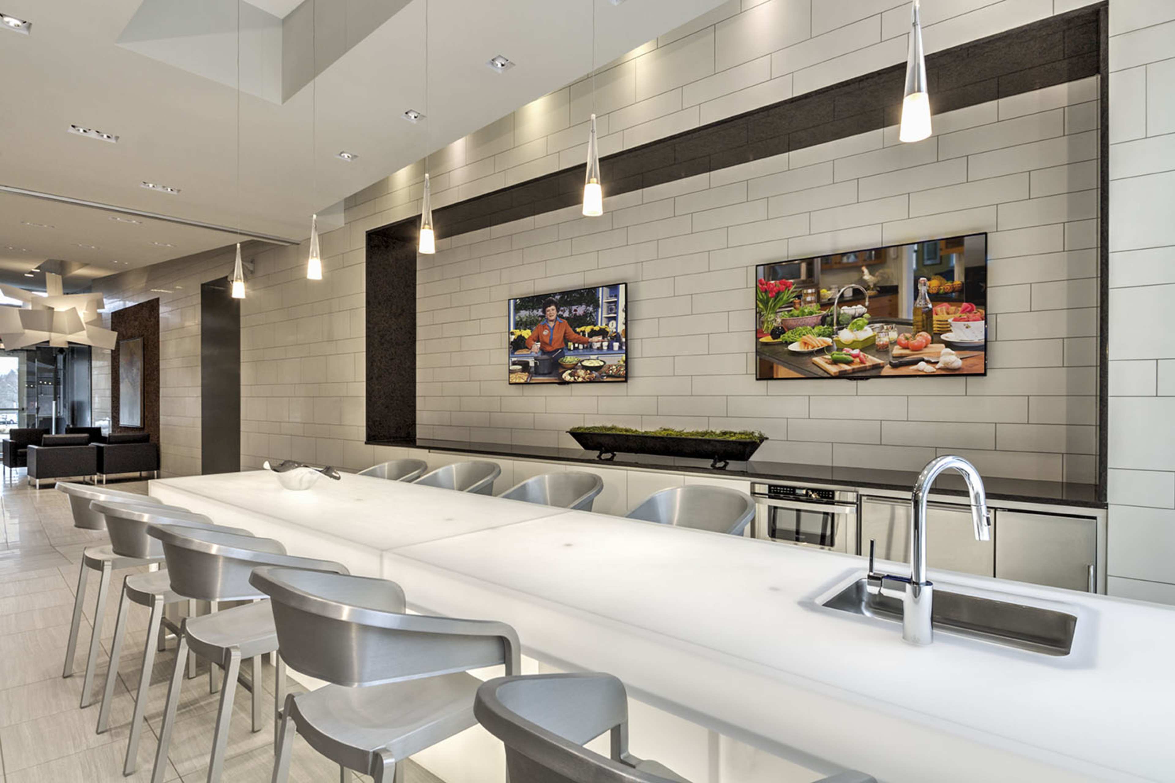A modern kitchen features a long white countertop with silver barstools, and two televisions on the wall displaying culinary images.