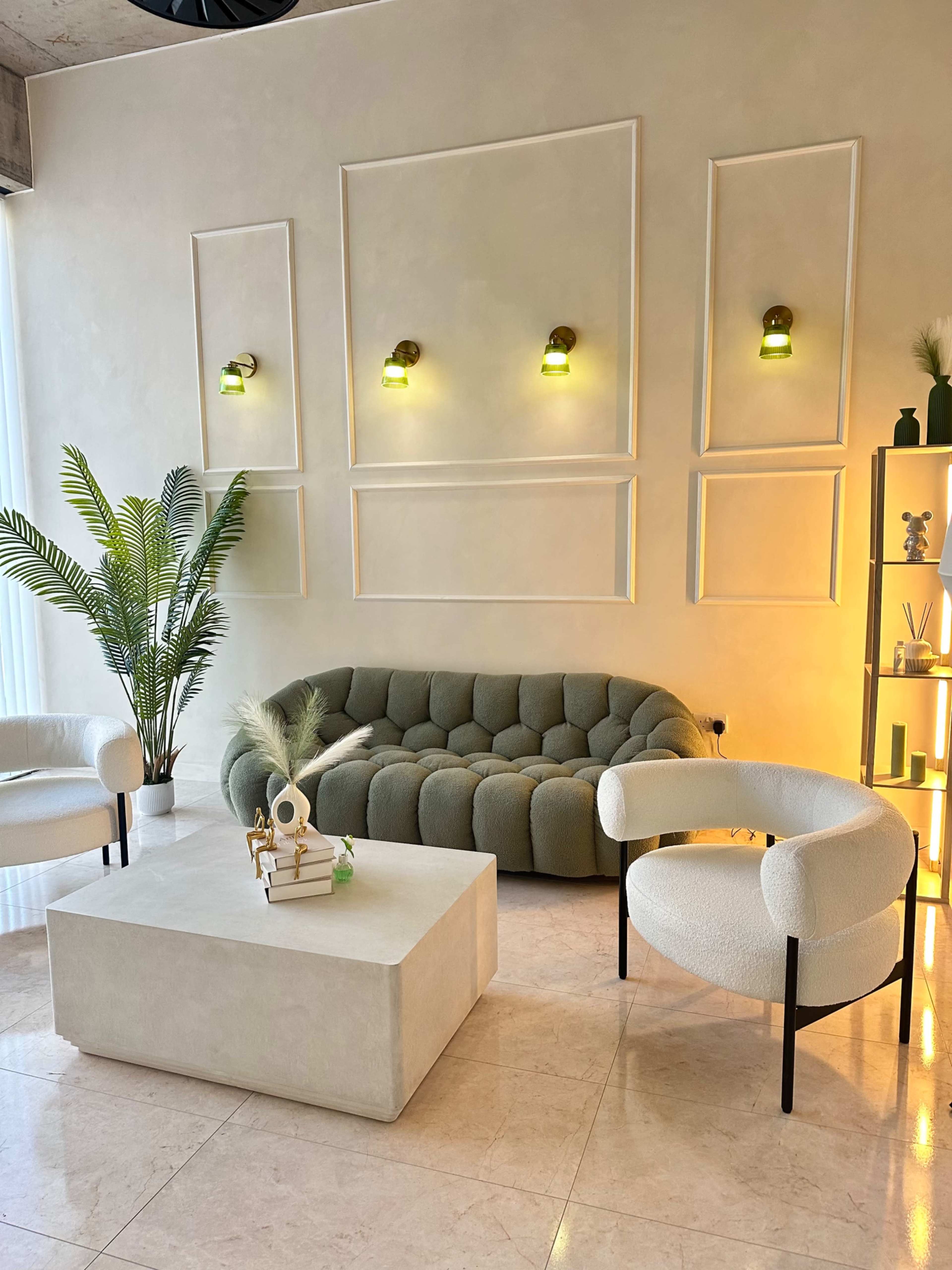 The image shows a modern living space featuring a rounded, green sofa, two white armchairs, a light-colored coffee table, and decorative shelving with plants and accents against a neutral wall.