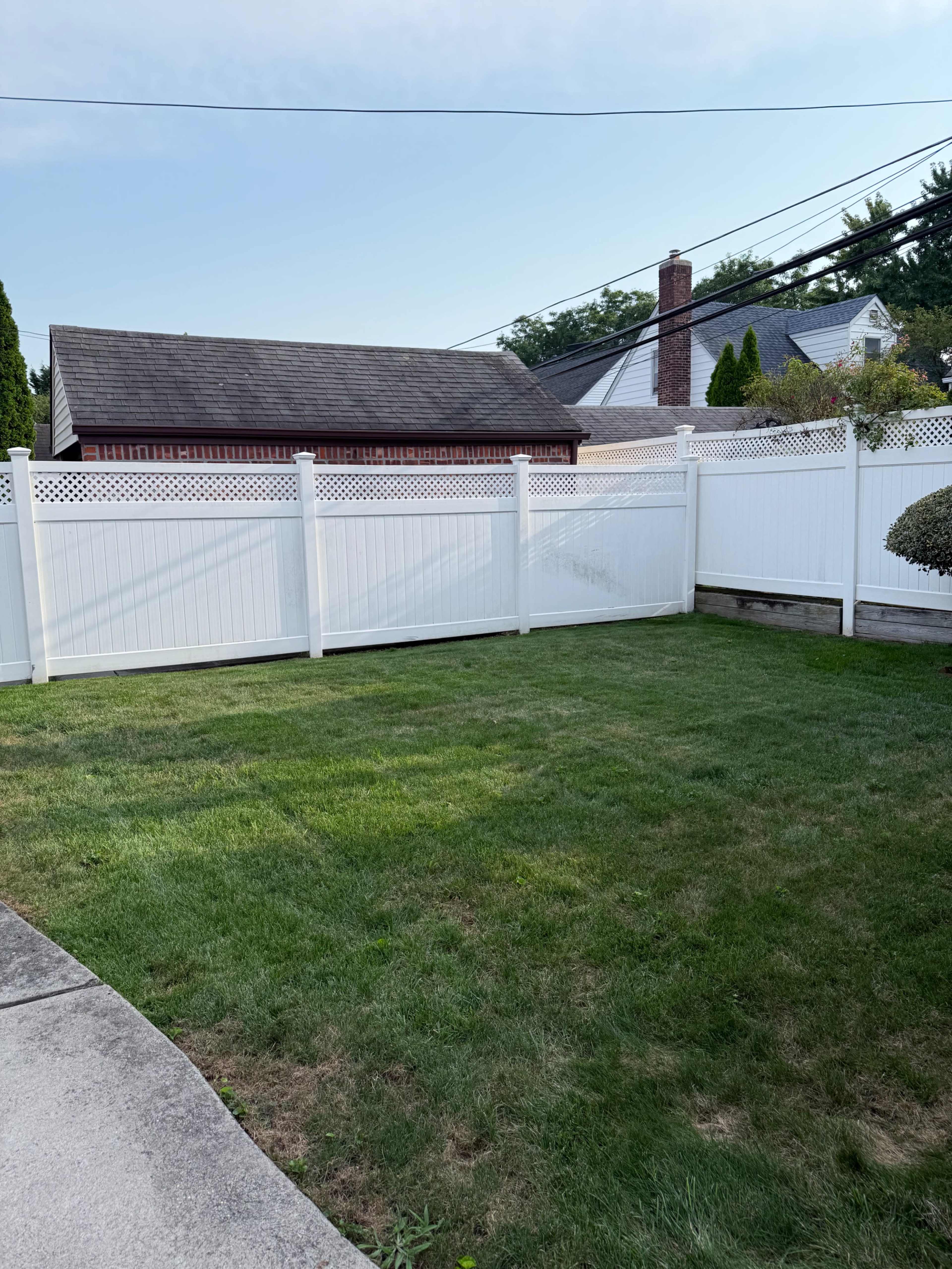 The image shows a well-maintained grassy backyard enclosed by a tall white fence with a brick building and utility poles visible in the background.