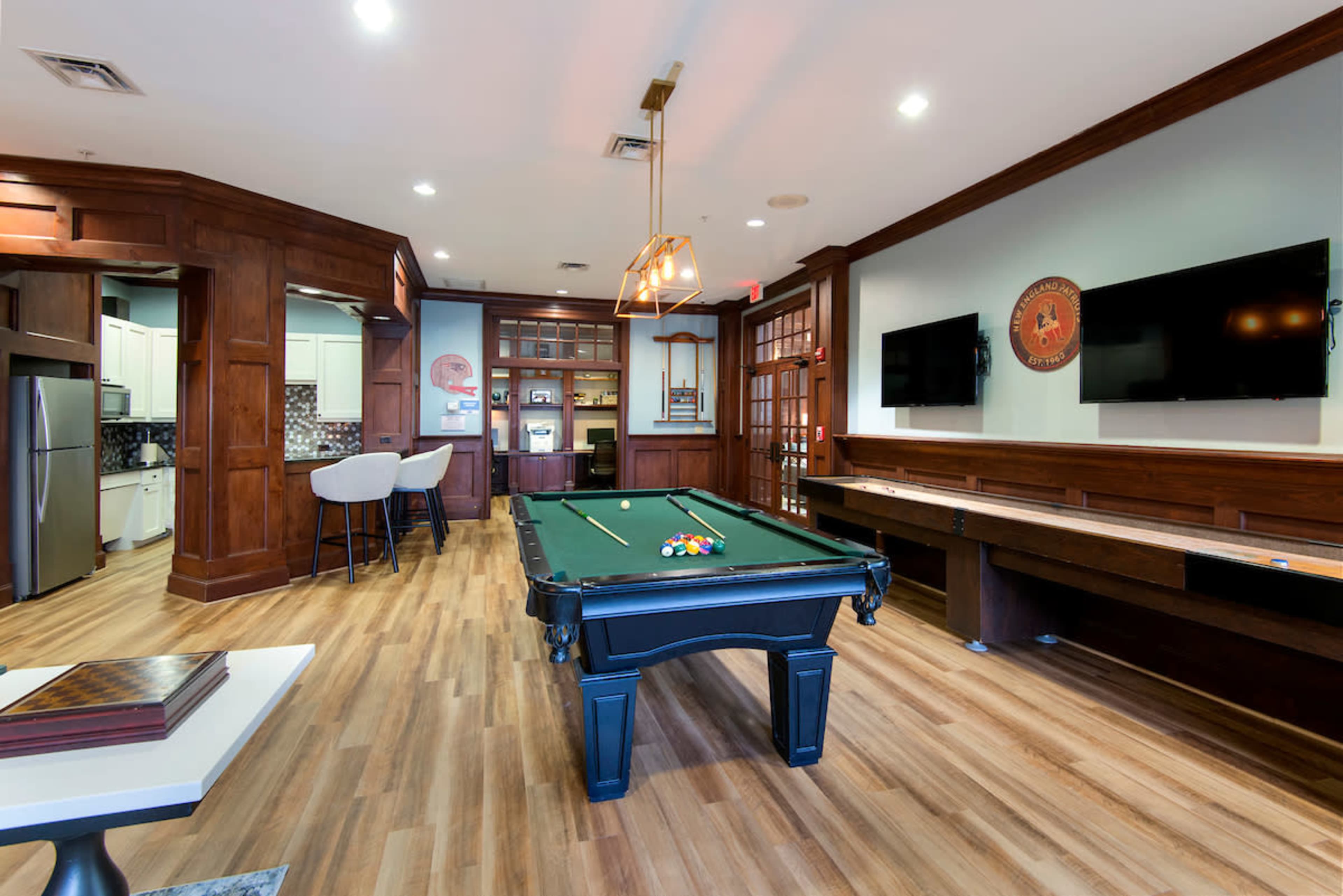 A pool table is centered in a game room featuring wooden paneling, a kitchenette area, and multiple televisions mounted on the walls.
