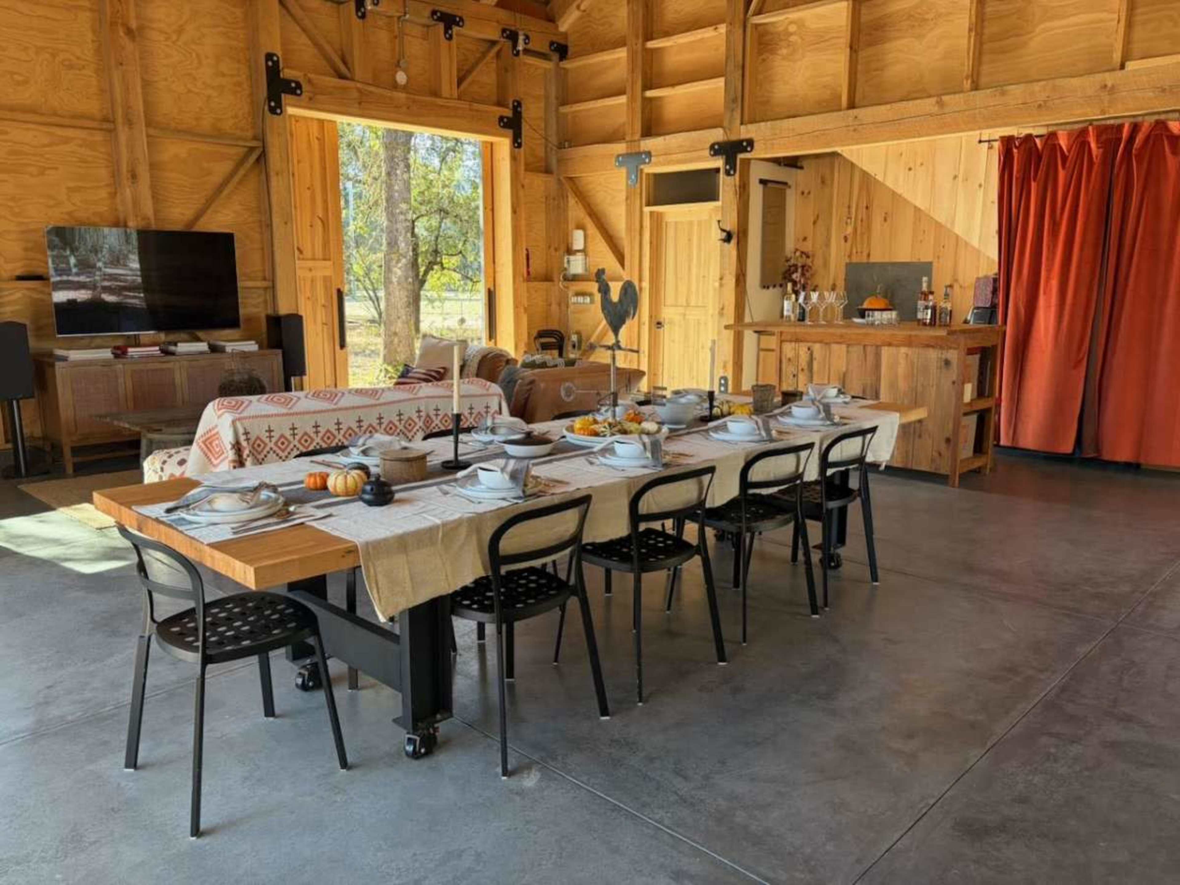 A spacious dining area features a long wooden table set for a meal, with a kitchen area and large windows letting in natural light.