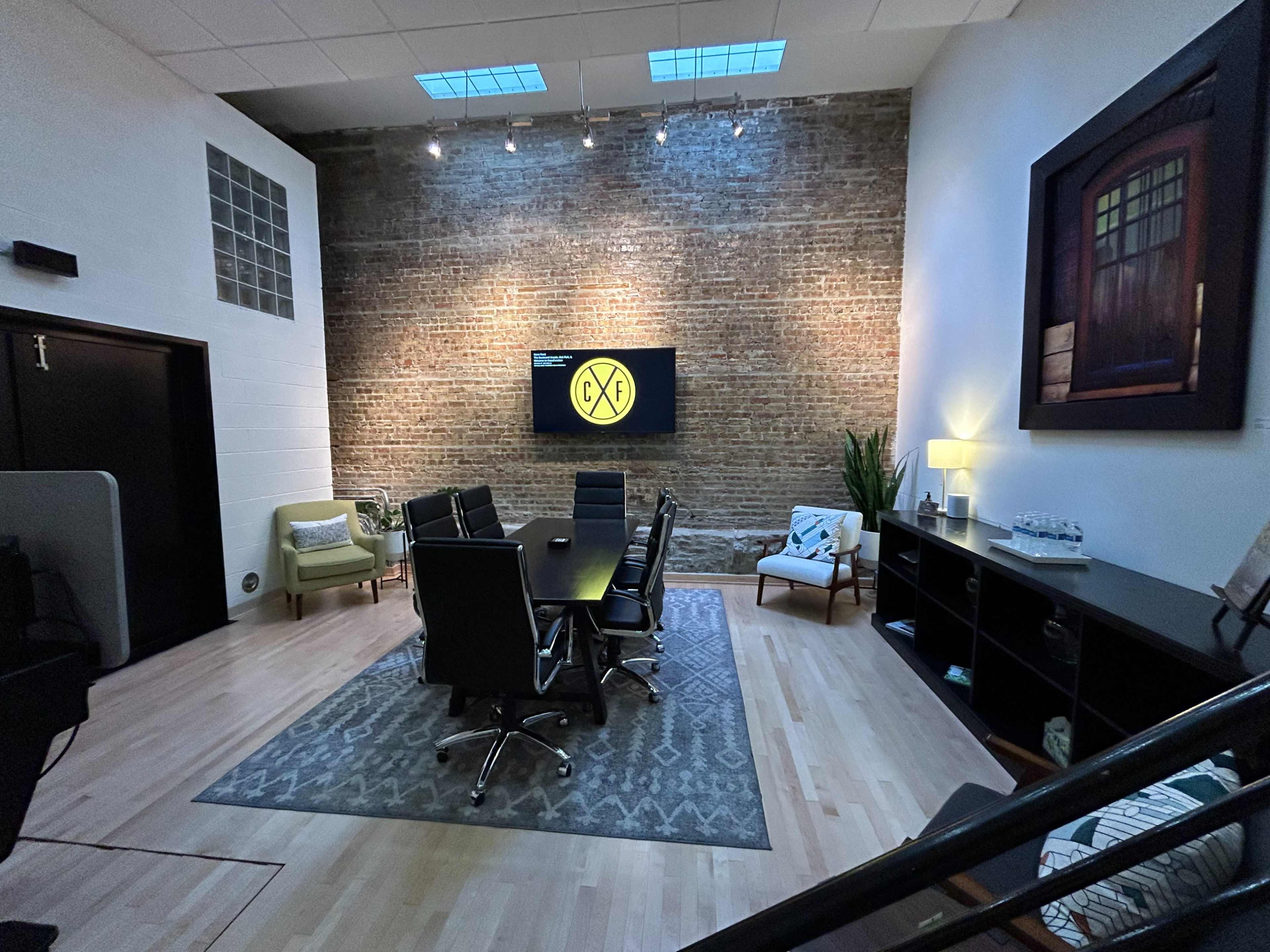 A modern conference room with a long table, black chairs, and a brick wall featuring a large screen displaying a logo.