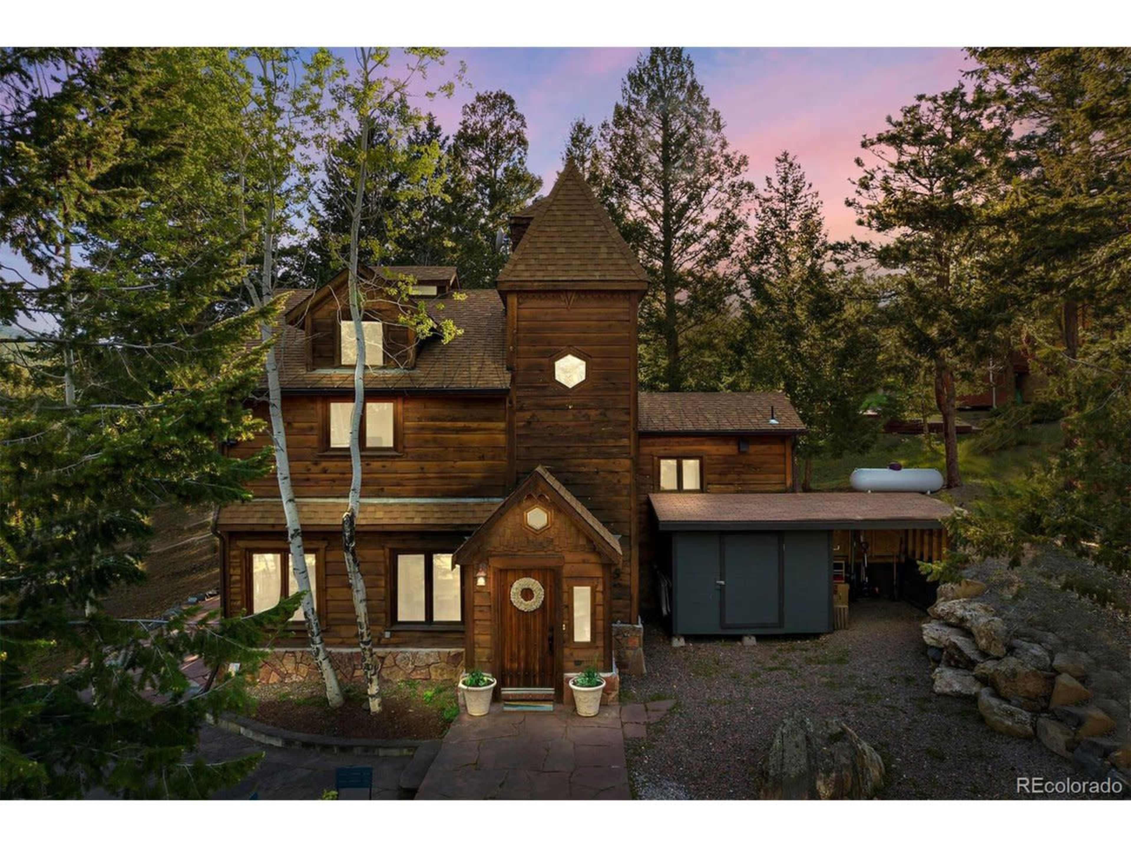 A multi-story wooden house surrounded by trees, featuring a unique tower and a stone pathway leading to the entrance.