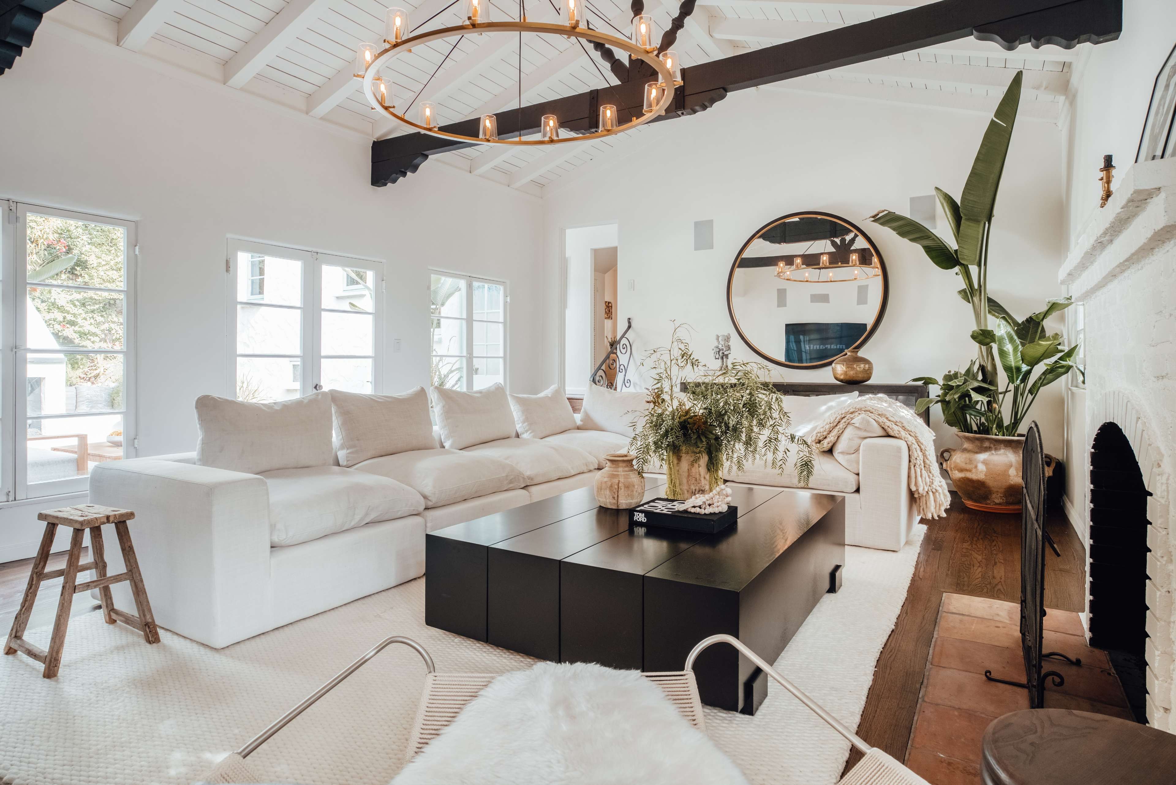 The image shows a spacious living room with a large sectional sofa, a dark coffee table, and a decorative mirror, accented by indoor plants and a fireplace.