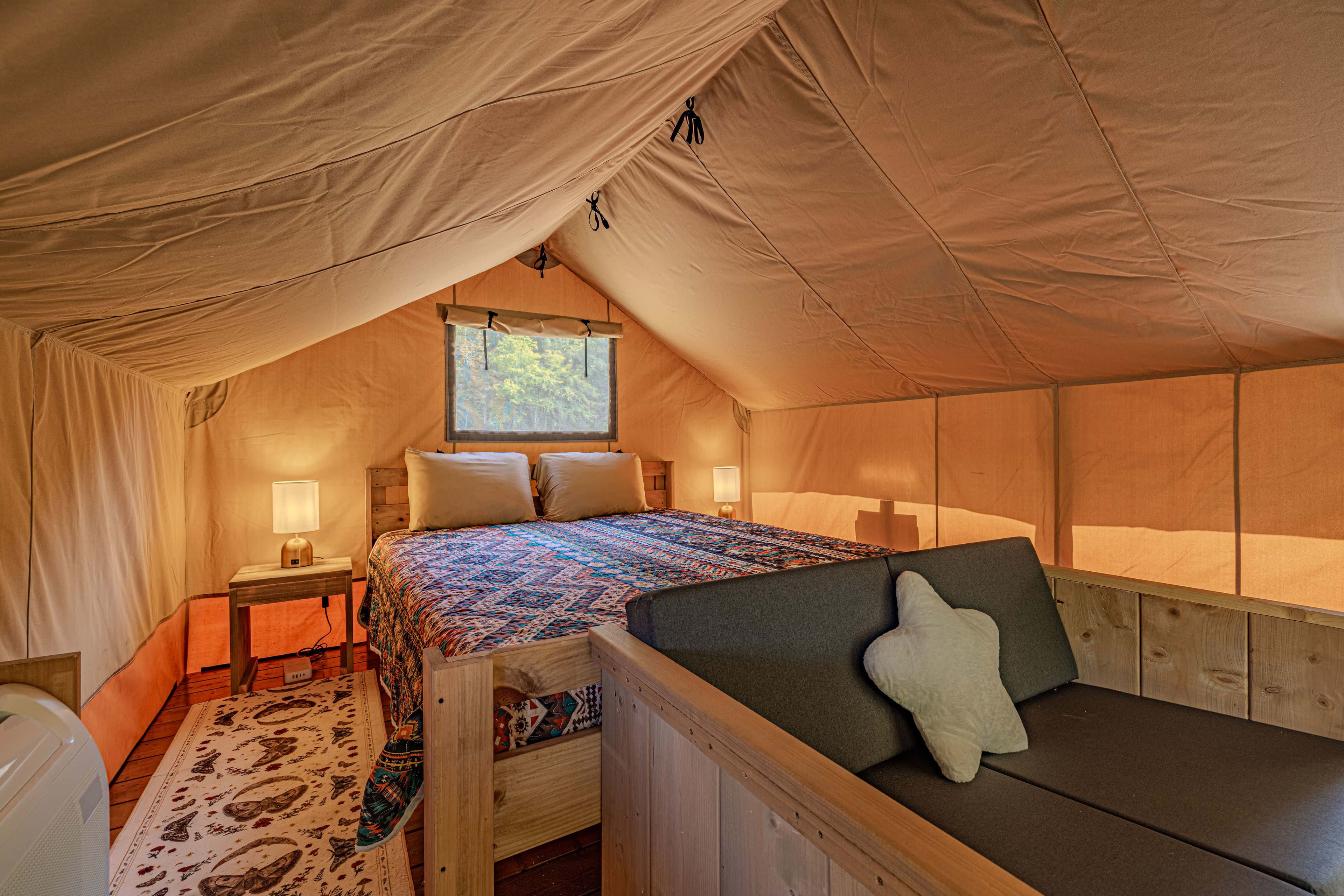 The image shows the interior of a cozy glamping tent featuring a large bed, a small table with lamps, and a seating area.