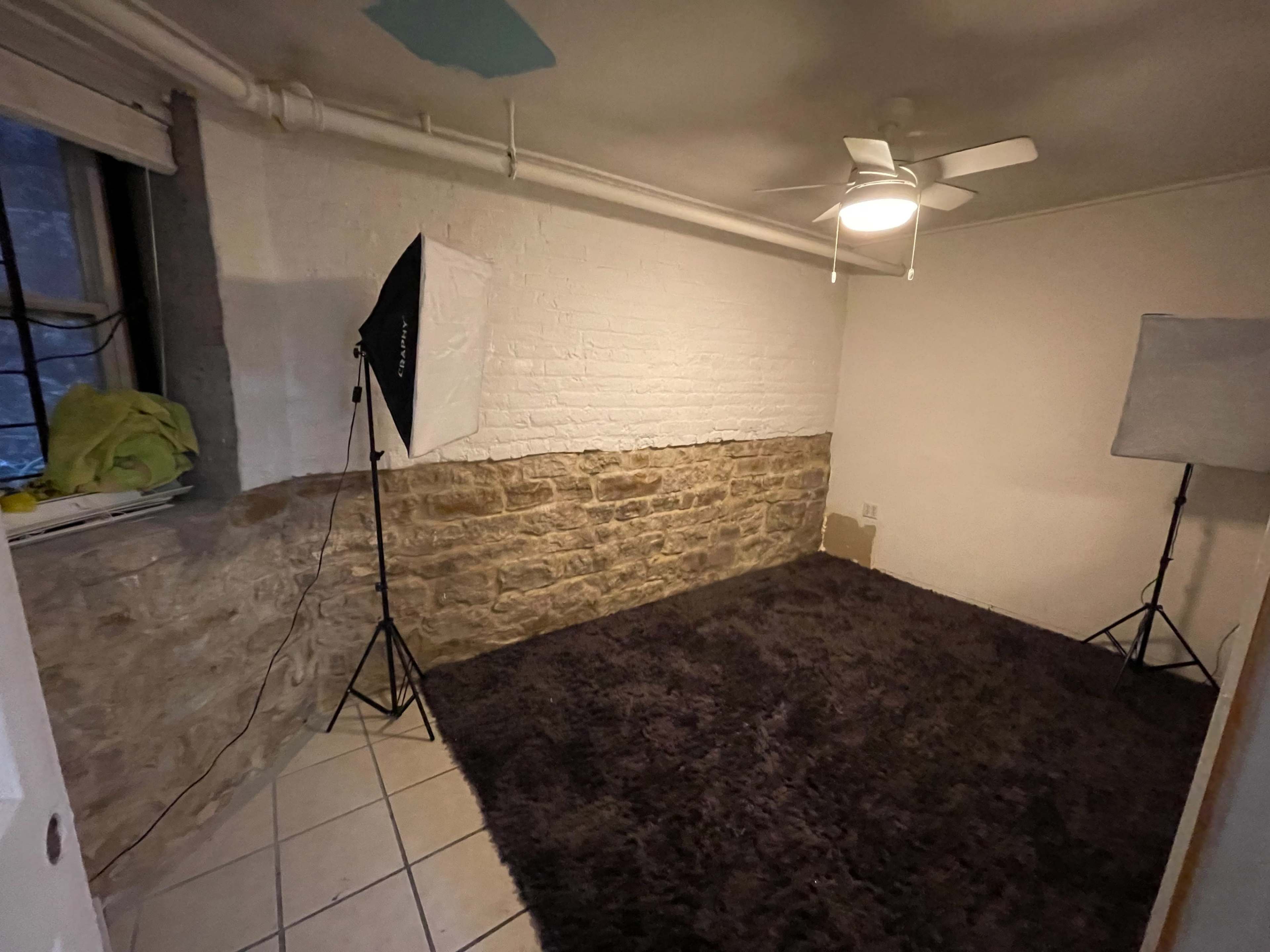 A sparsely furnished room with a stone wall on one side, a ceiling fan, and photography lights set up in the corners.