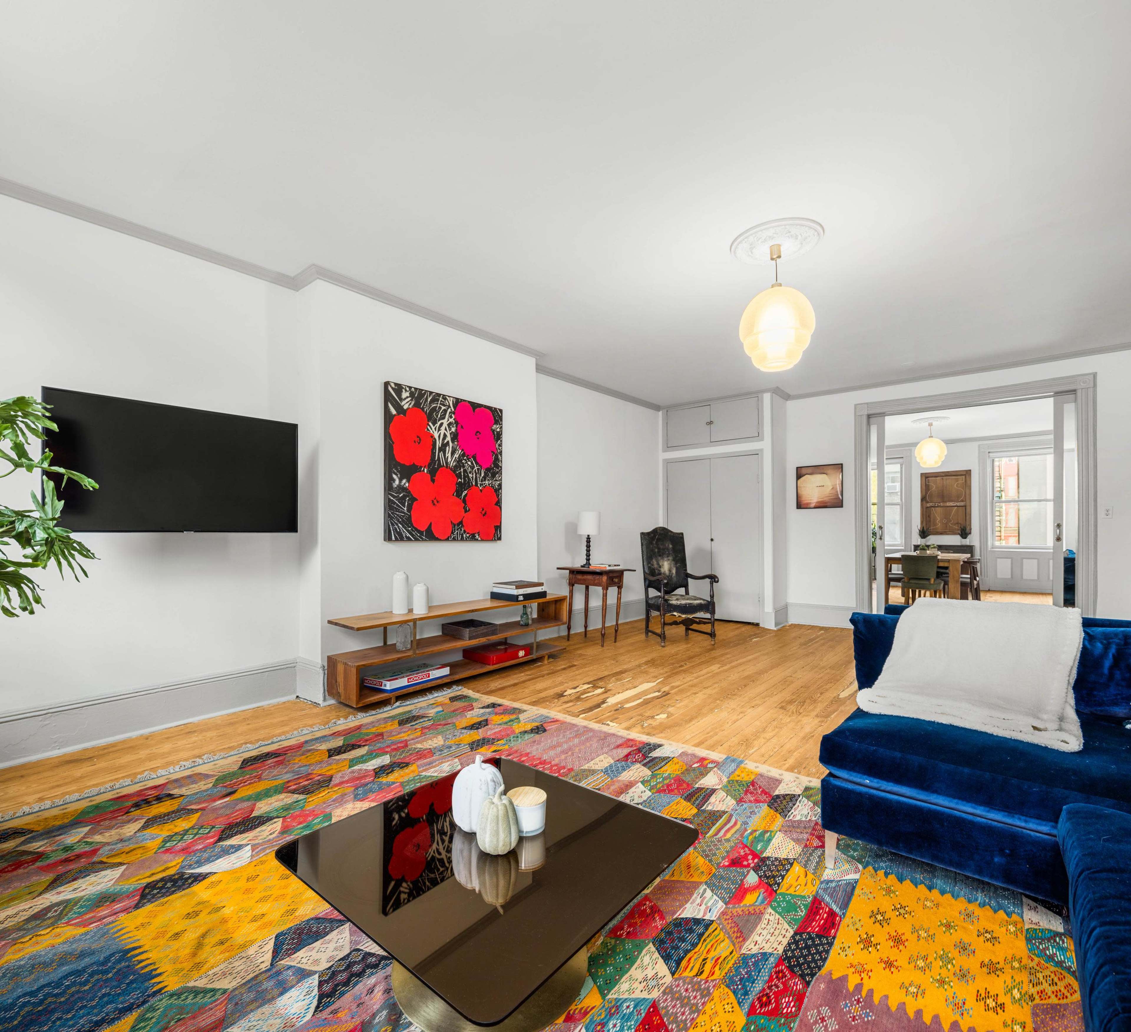 A living room features a colorful patterned rug, a blue sofa, a coffee table, and a wall-mounted television, with an artwork and a potted plant in the background.
