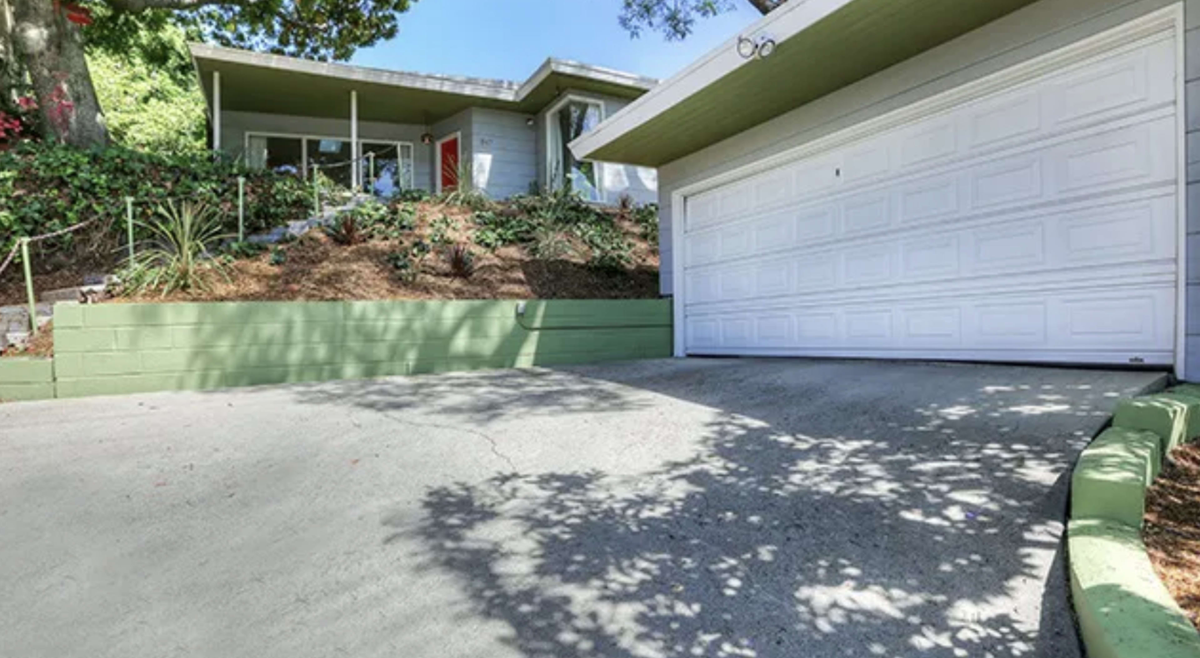 A driveway leading up to a house with a gray exterior and a white garage door, surrounded by greenery.