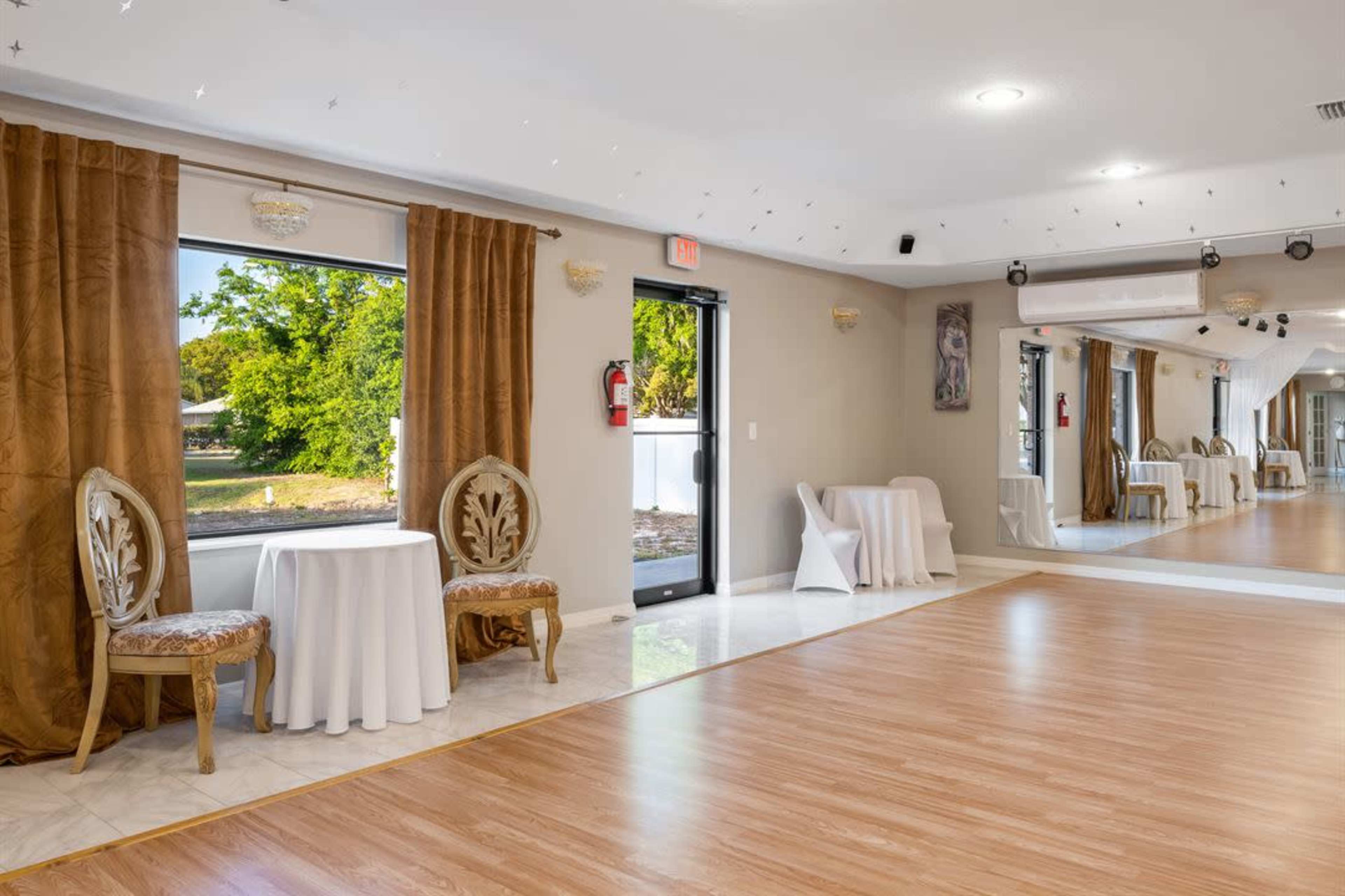 The photo shows a spacious room with wooden floors, two ornate chairs near a small table, and large windows covered with brown curtains.