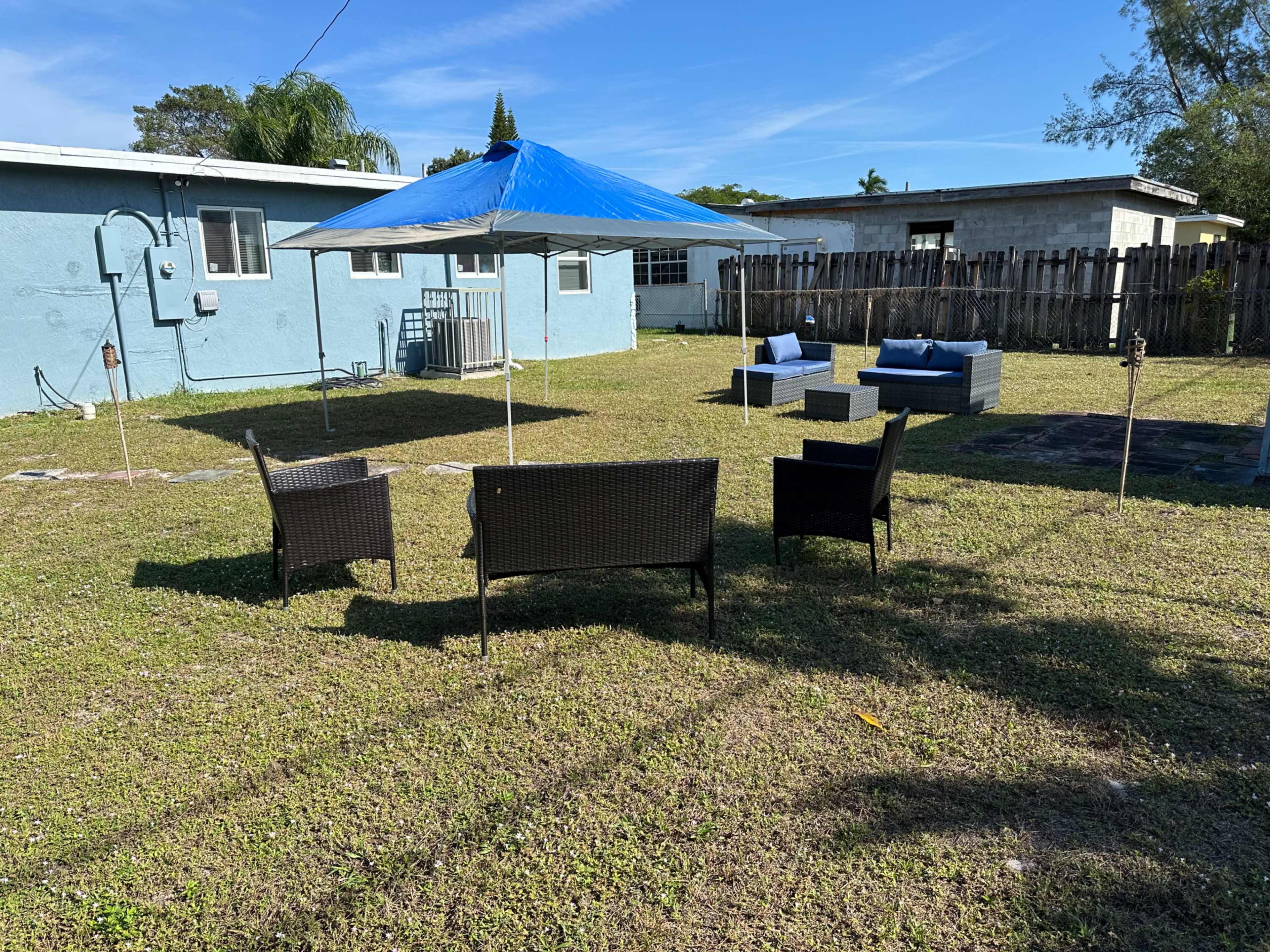 The image shows a grassy backyard with four patio chairs under a blue umbrella and a sectional outdoor sofa in the background.