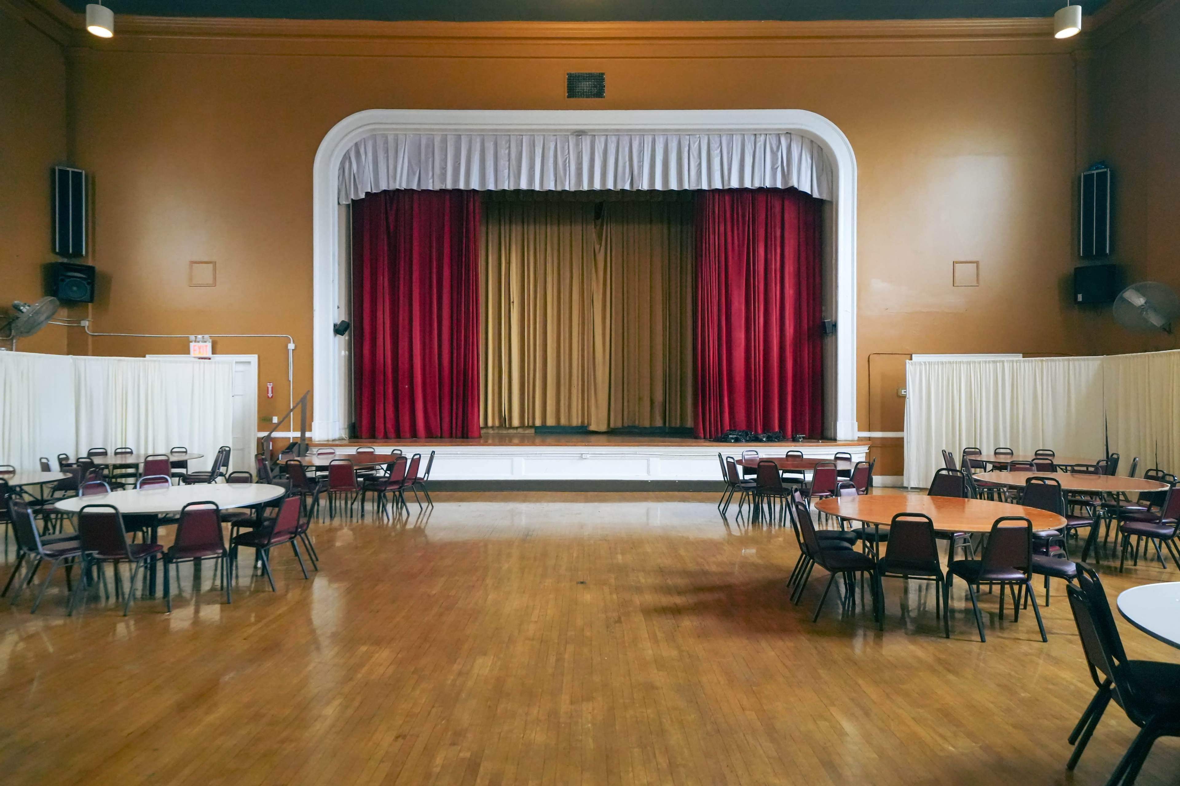 A large hall with several round tables arranged in front of a stage draped in red and gold curtains.