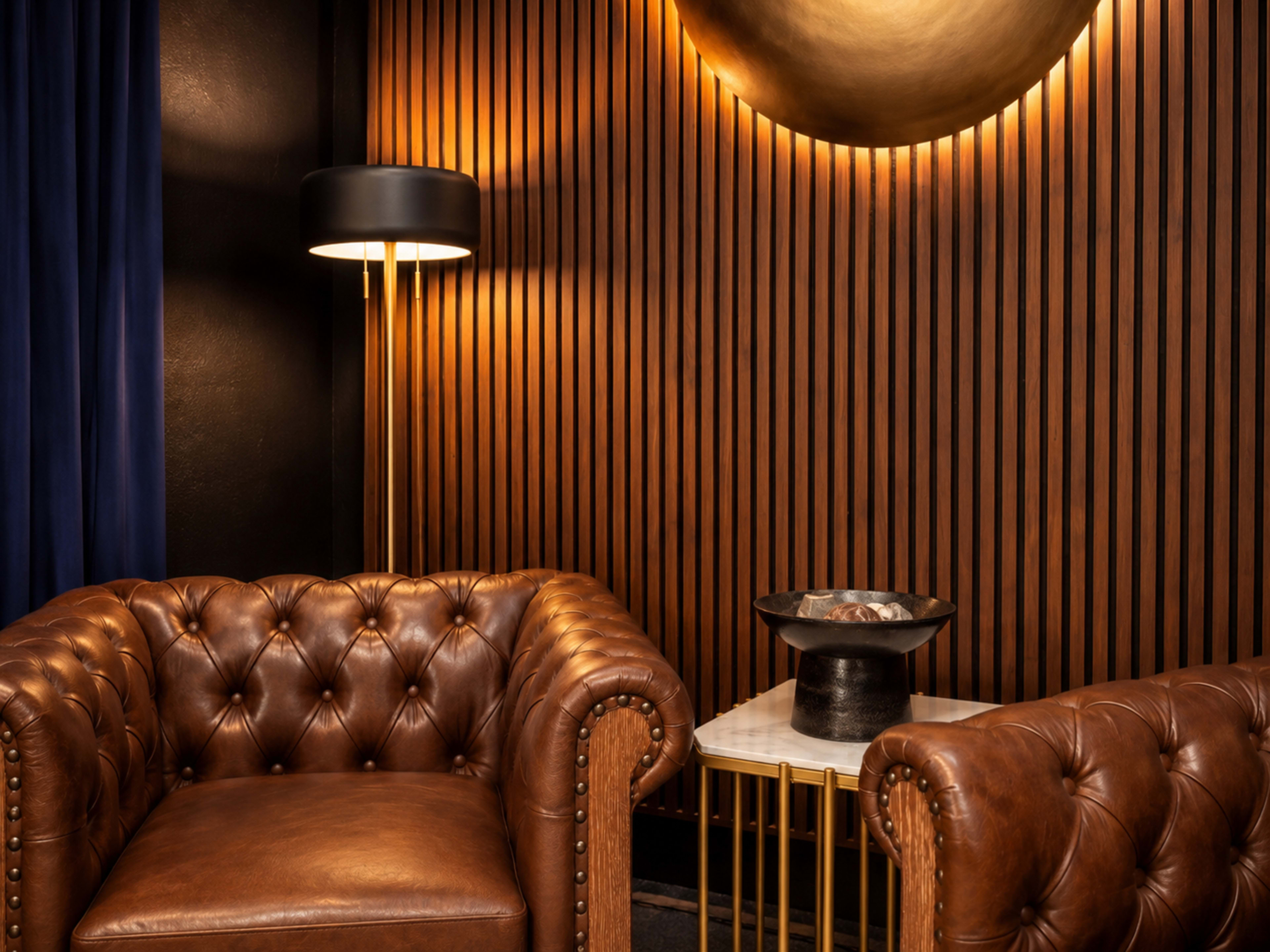 The image shows a cozy corner featuring two brown leather armchairs, a marble-topped side table, and a modern lamp against a backdrop of wooden paneling.