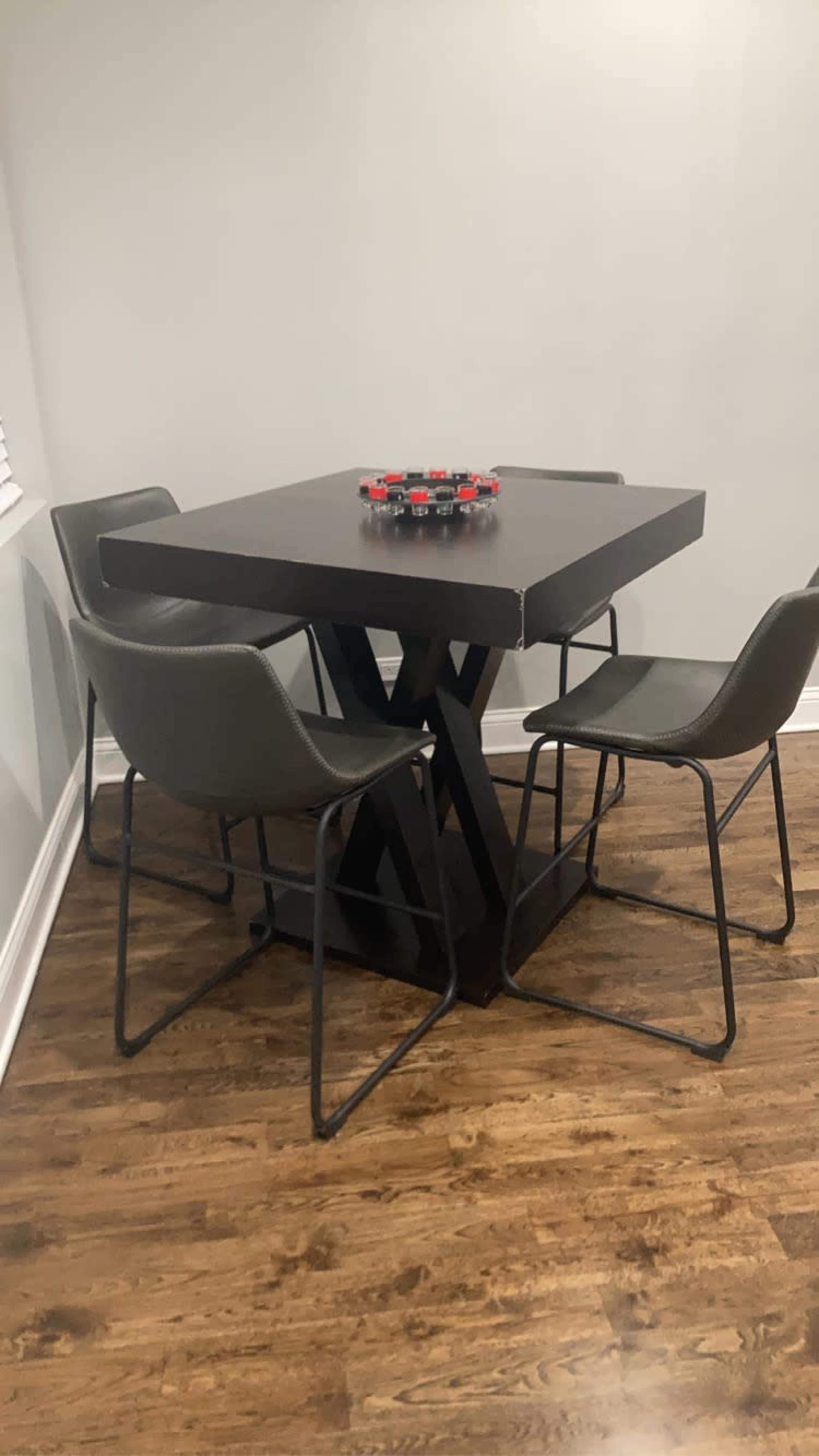 A square wooden table is positioned in a room with four green chairs around it, and a circular arrangement of small items sits on the table's surface.