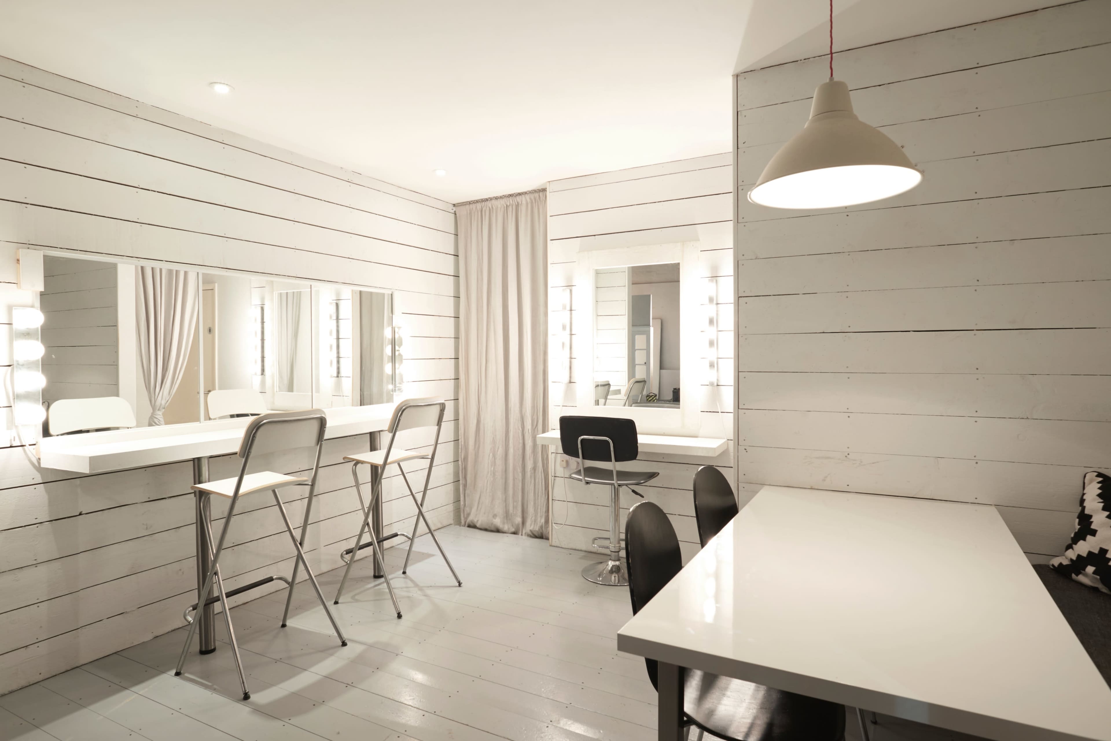 A minimalist dressing room with white wooden walls, two large mirrors framed by lights, and a variety of seating options.