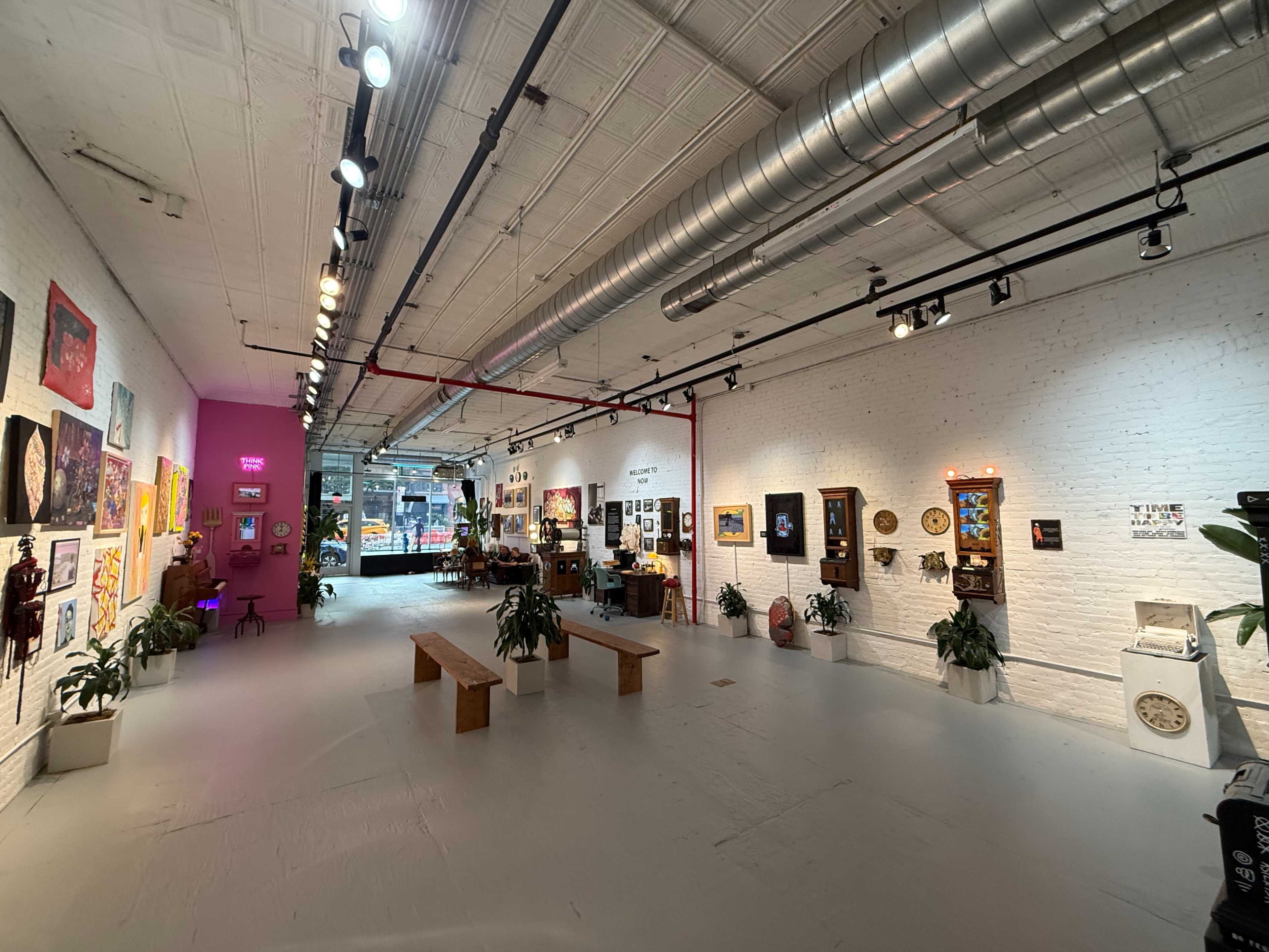 The image shows an art gallery with white walls displaying various artworks, wooden benches, and plants scattered throughout the space.