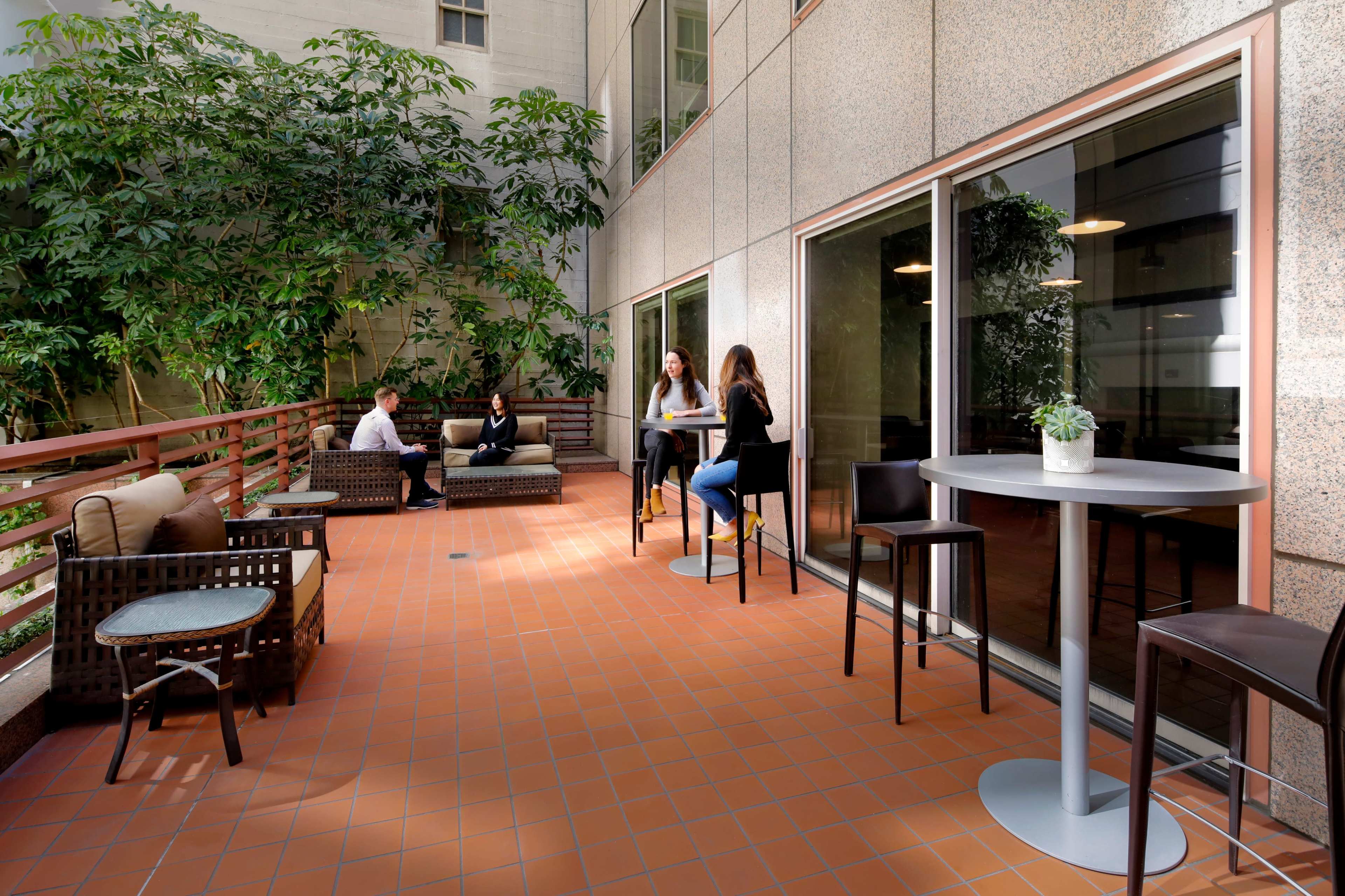 A small outdoor patio features people seated at tables and relaxing on couches surrounded by lush greenery.