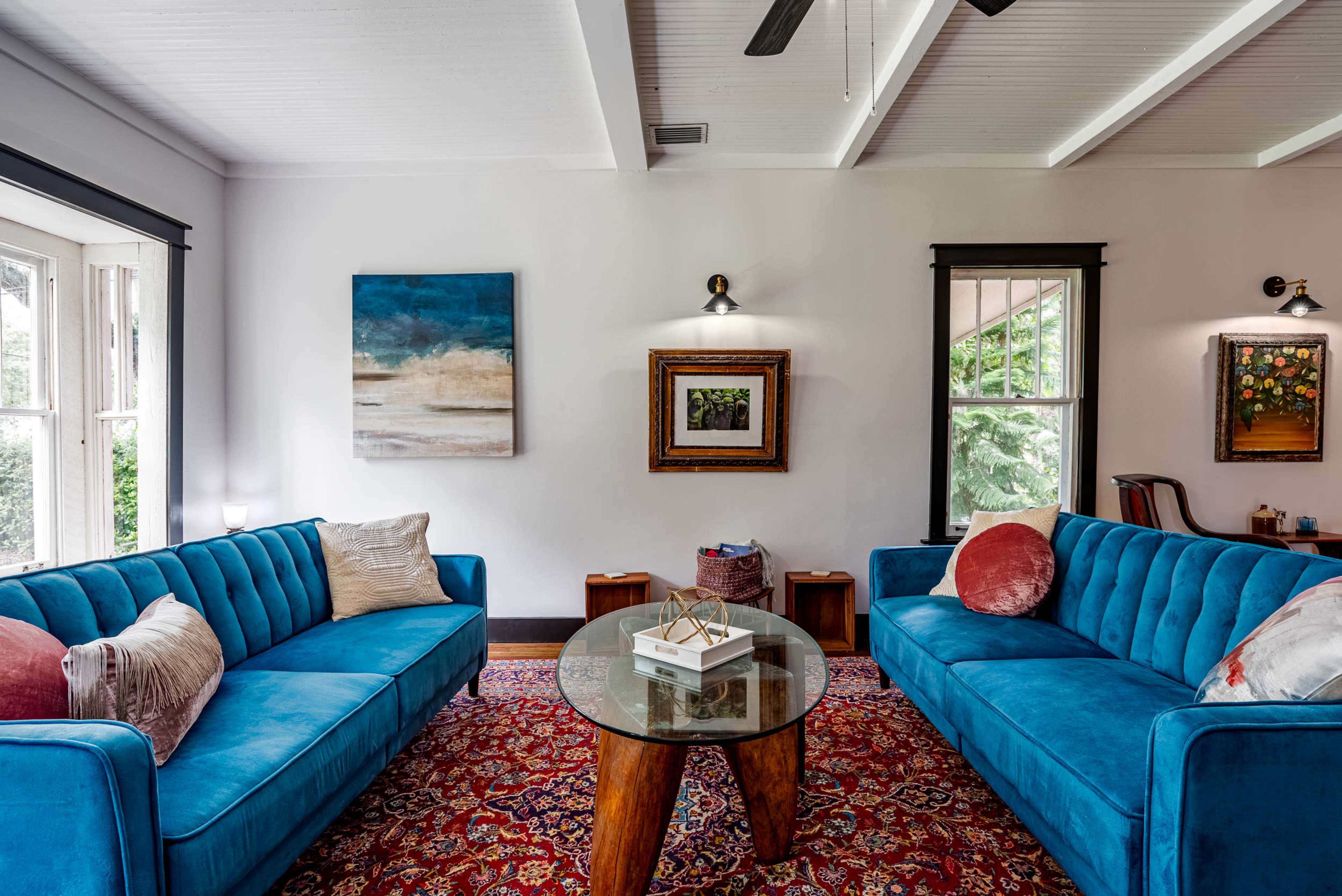 A living room features two blue sofas facing a round glass table on a patterned rug, with artwork and windows allowing natural light.