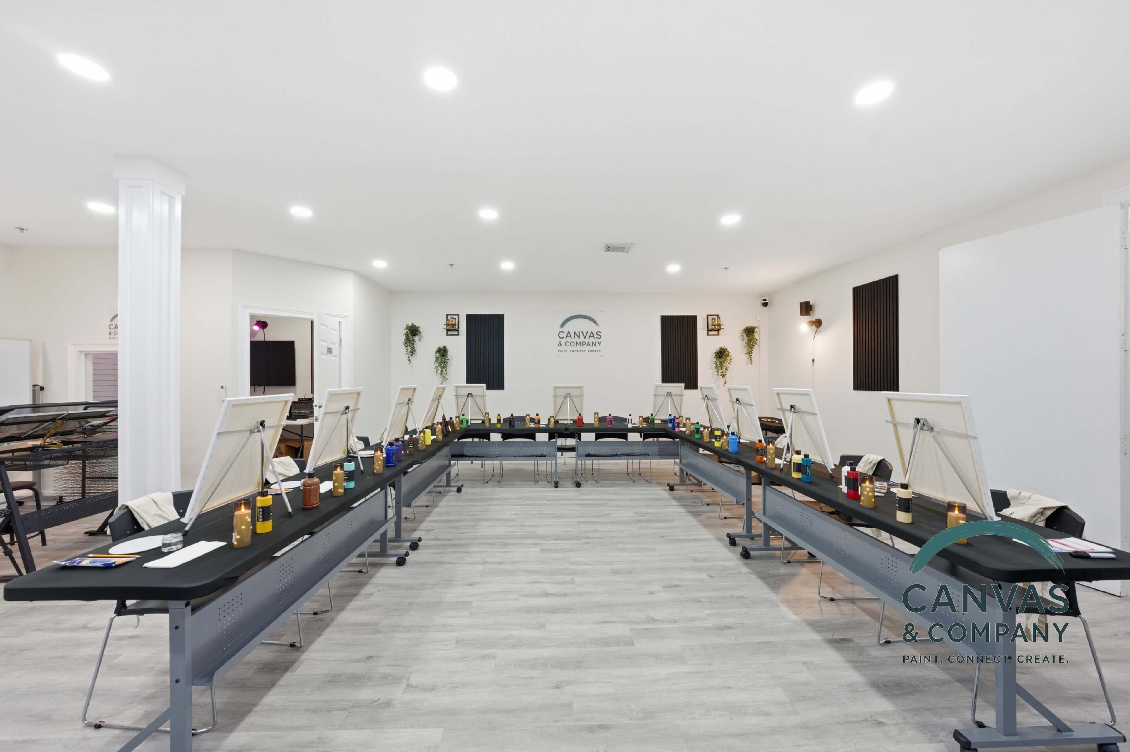 The image shows a spacious art studio with several painting stations, each equipped with easels and paint supplies arranged neatly along long tables.