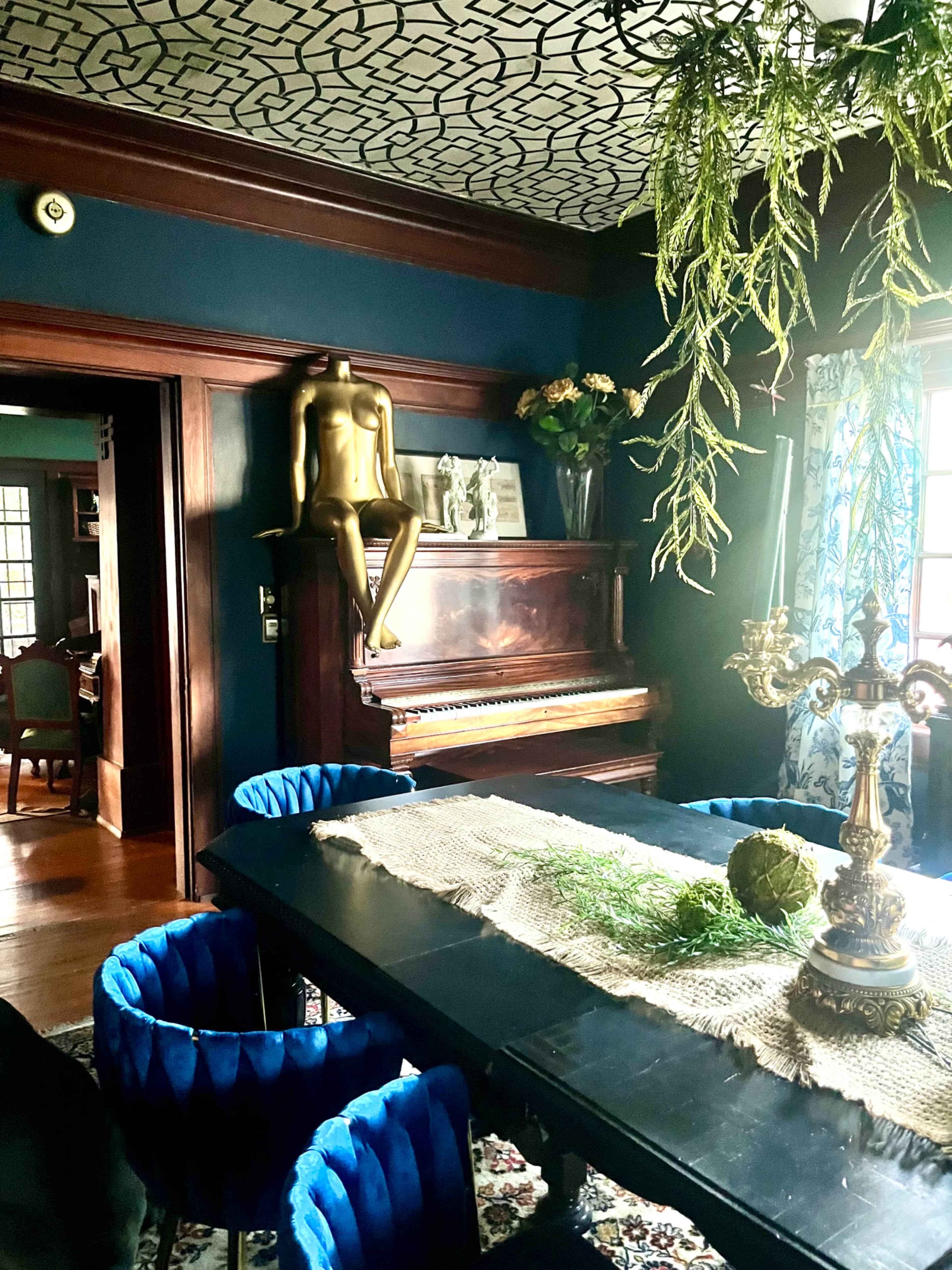 The image depicts a dining area featuring a dark blue wall, a wooden piano, a sculptural figure on the piano, a long black dining table with blue chairs, and decorative greenery hanging from the ceiling.