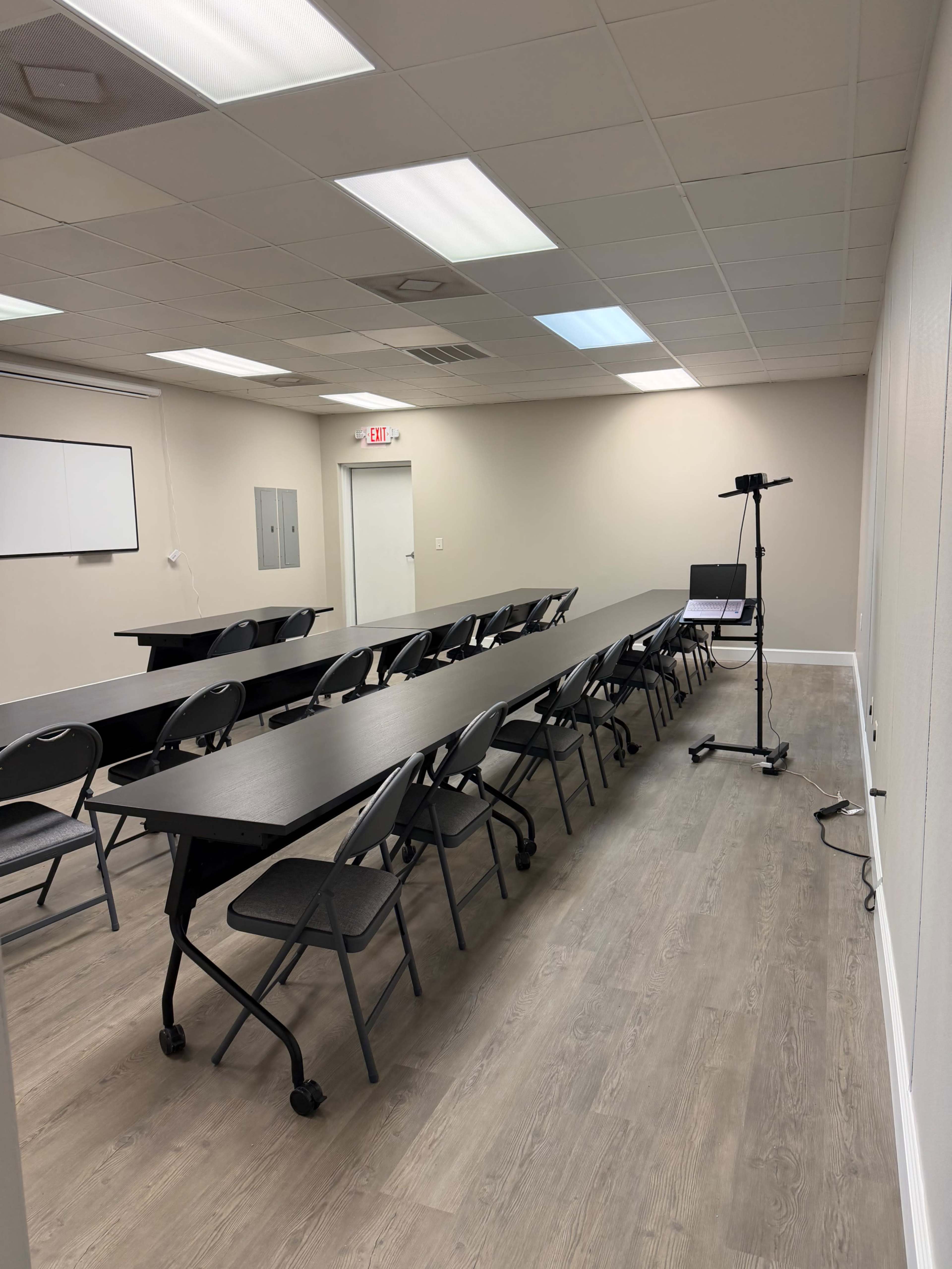 The image shows a sparsely furnished meeting room with several folding chairs arranged along long tables and a laptop stand in one corner.