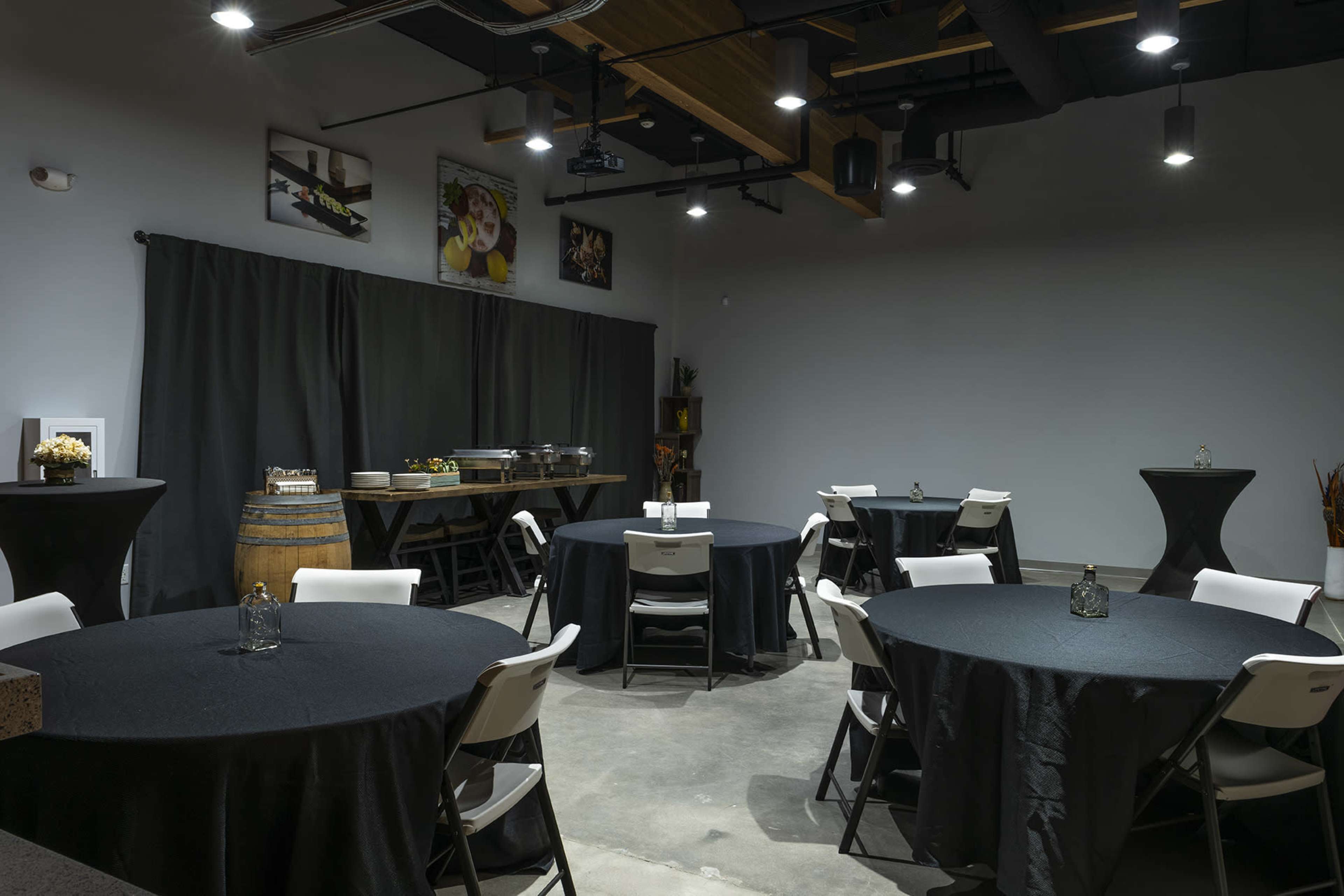 The image shows an interior space with several round tables covered in black tablecloths, a buffet setup in the background, and soft lighting fixtures.