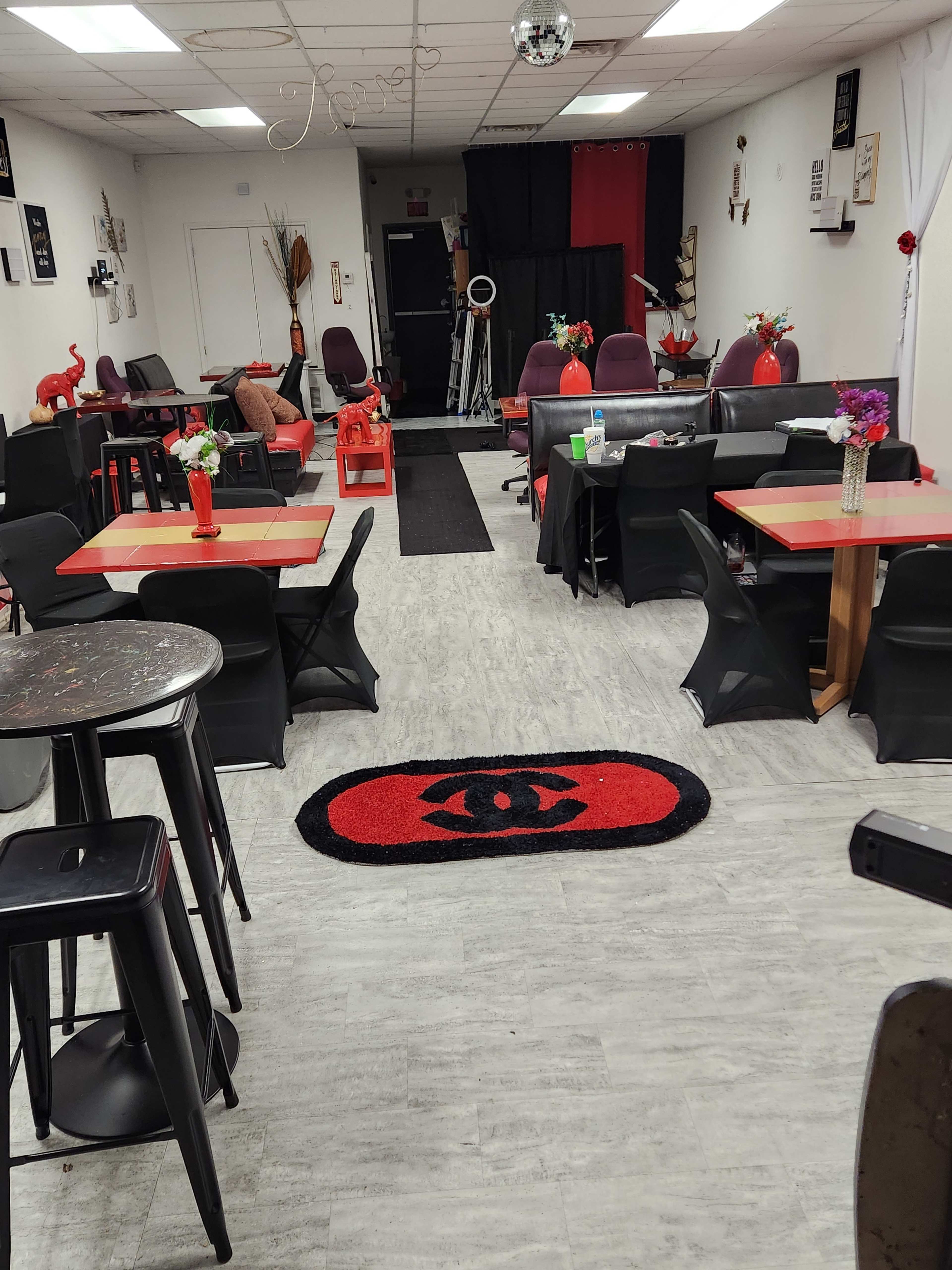 An indoor café or lounge area with red and black décor, featuring tables, chairs, and a lengthy black carpet running down the center.