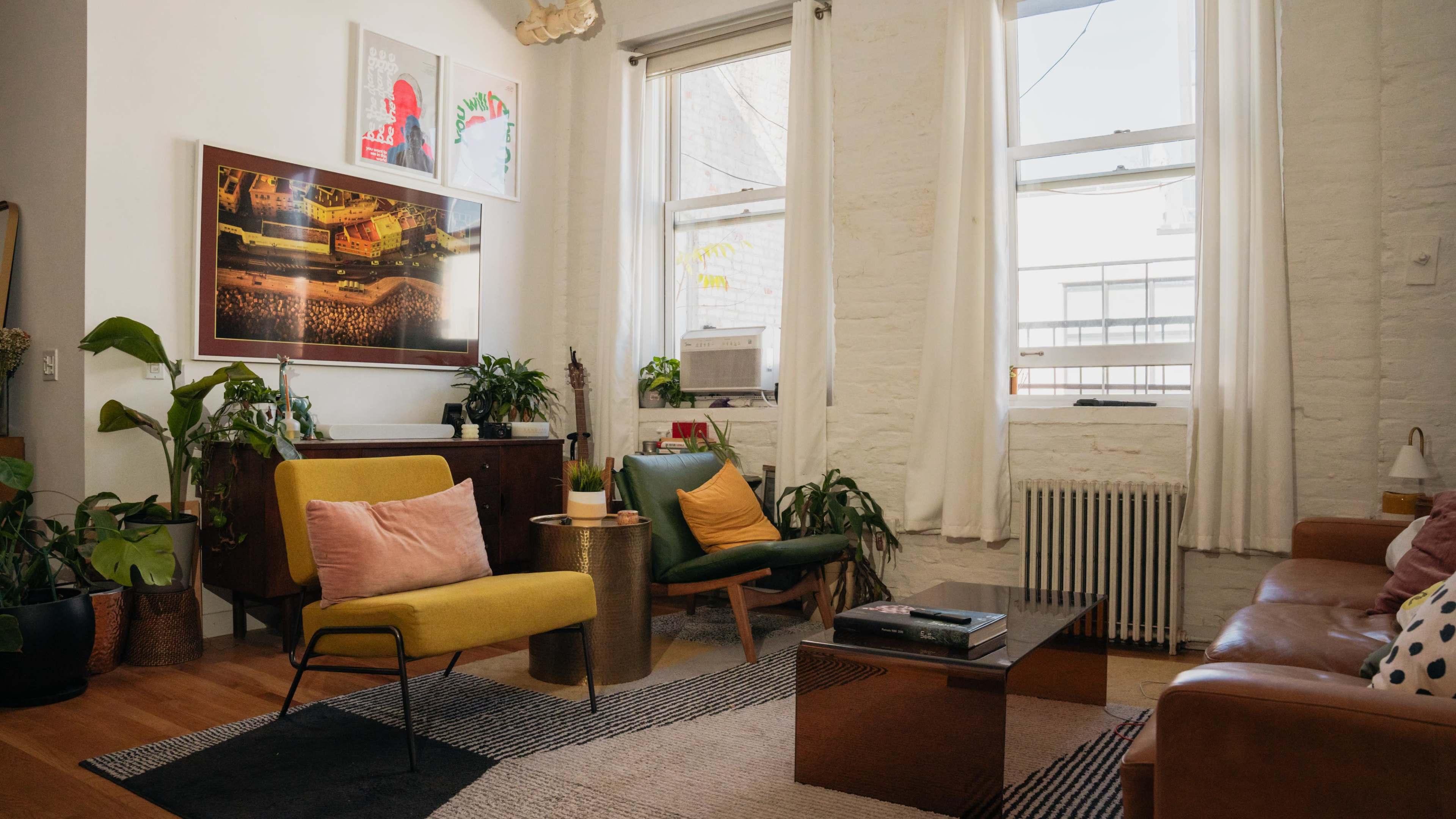 A bright living room features green and yellow chairs, a wooden coffee table, and various indoor plants against a backdrop of white brick walls.