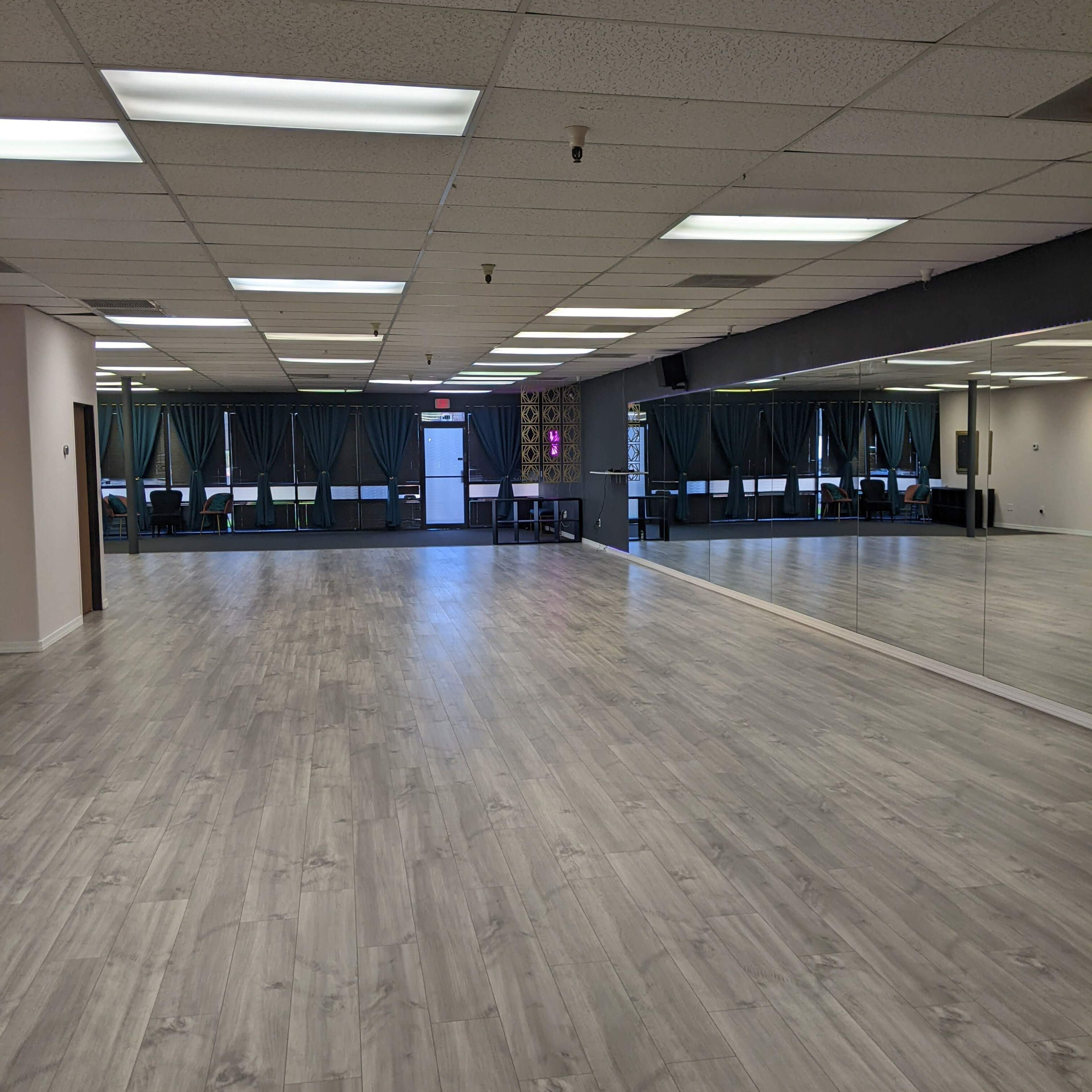 A spacious dance studio with mirrored walls, gray flooring, and large windows covered by teal curtains.