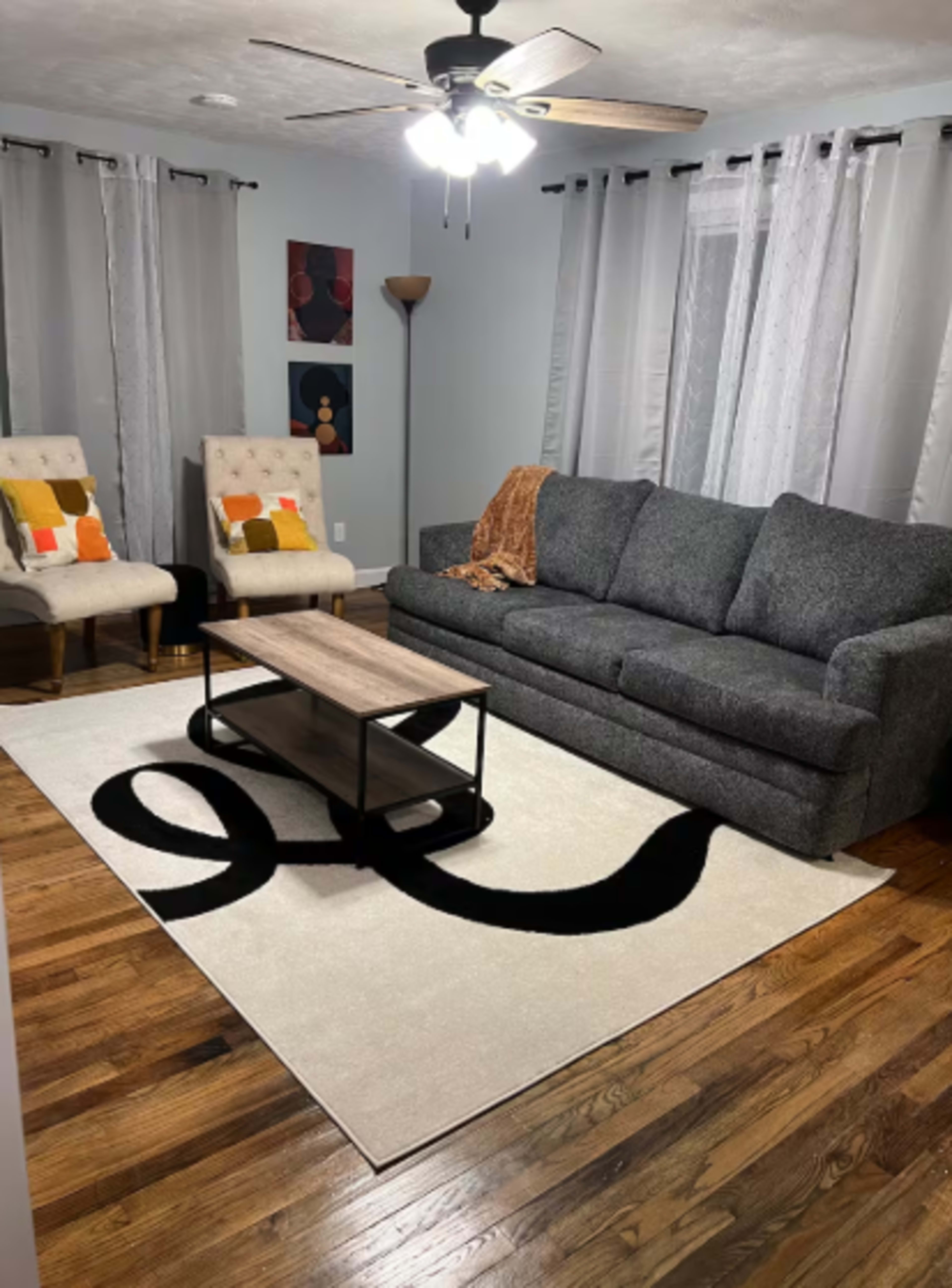 The image shows a living room featuring a gray couch, two beige chairs with colorful cushions, a coffee table, and a light-colored rug with a black design on a hardwood floor.